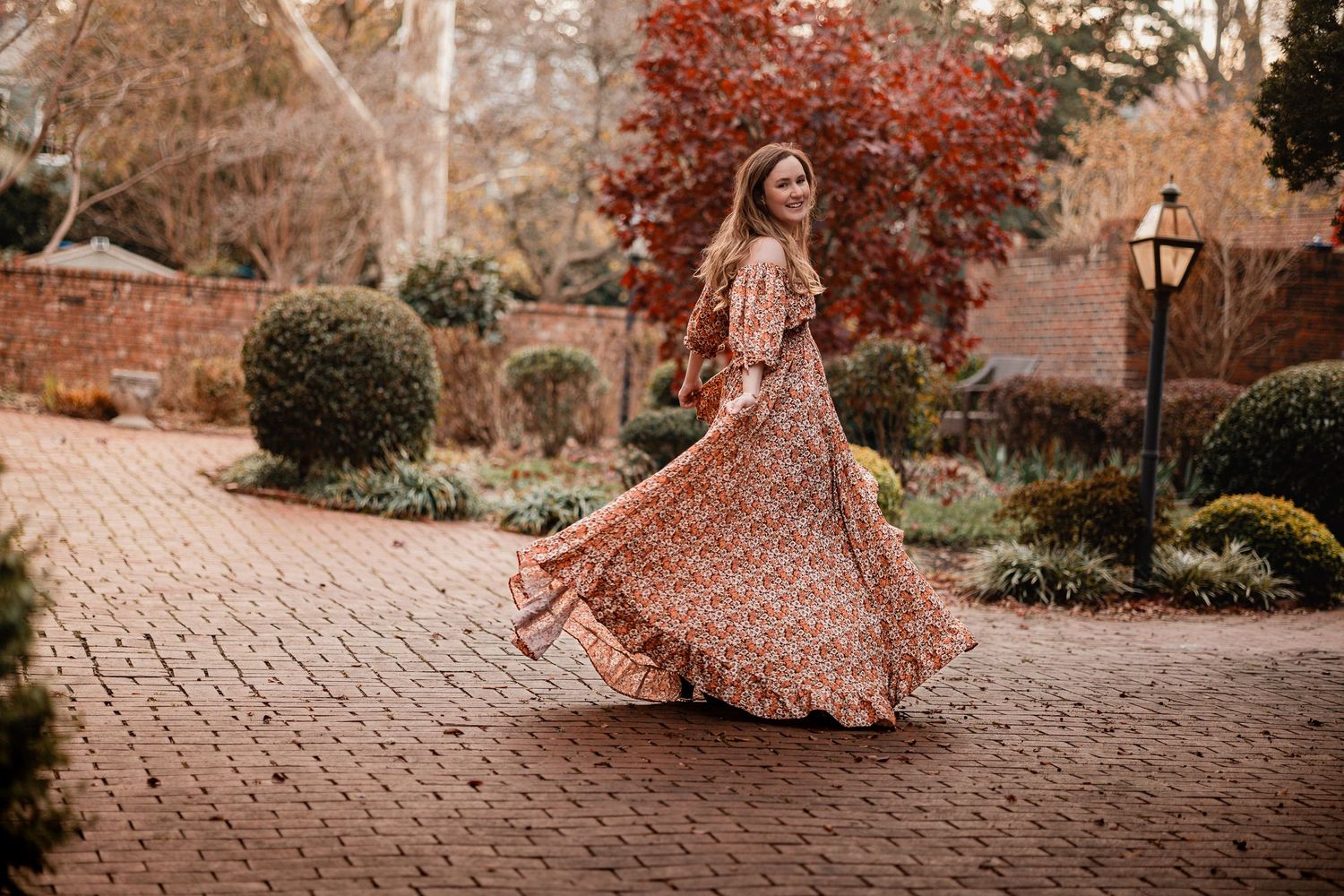 A person in a flowing rose gold sequined gown twirls on a cobblestone path surrounded by autumn foliage.