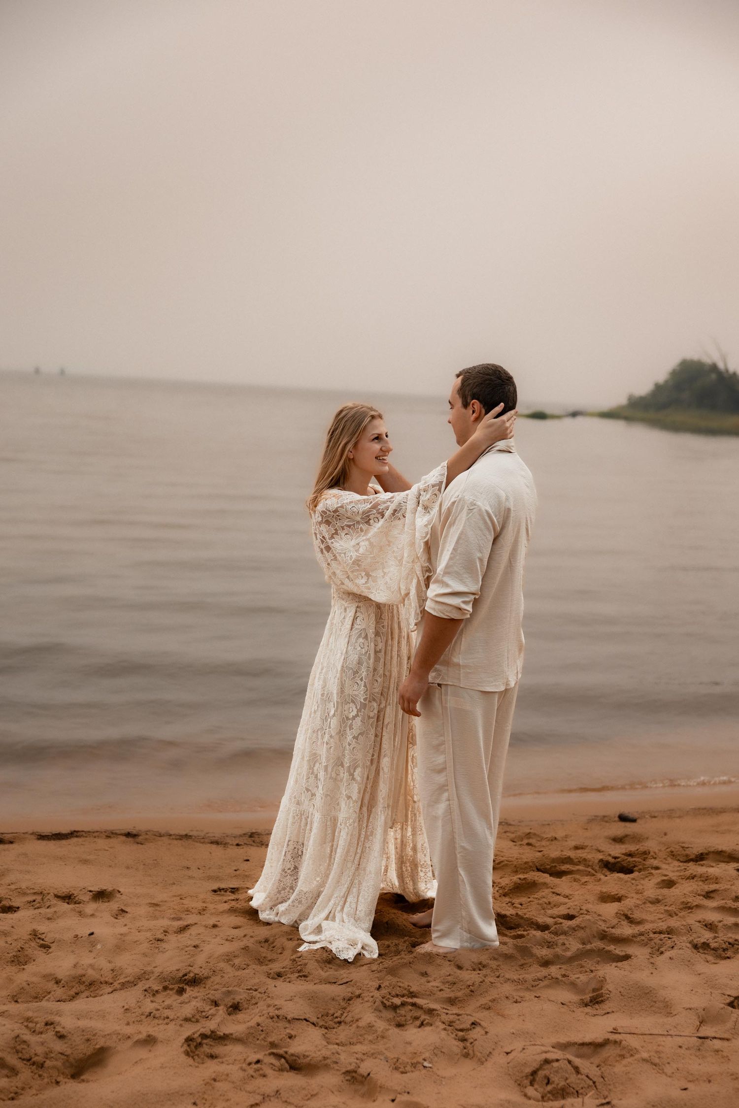 Couple in white attire embracing on sandy beach at dusk, woman in lace dress touching mans face by calm water.