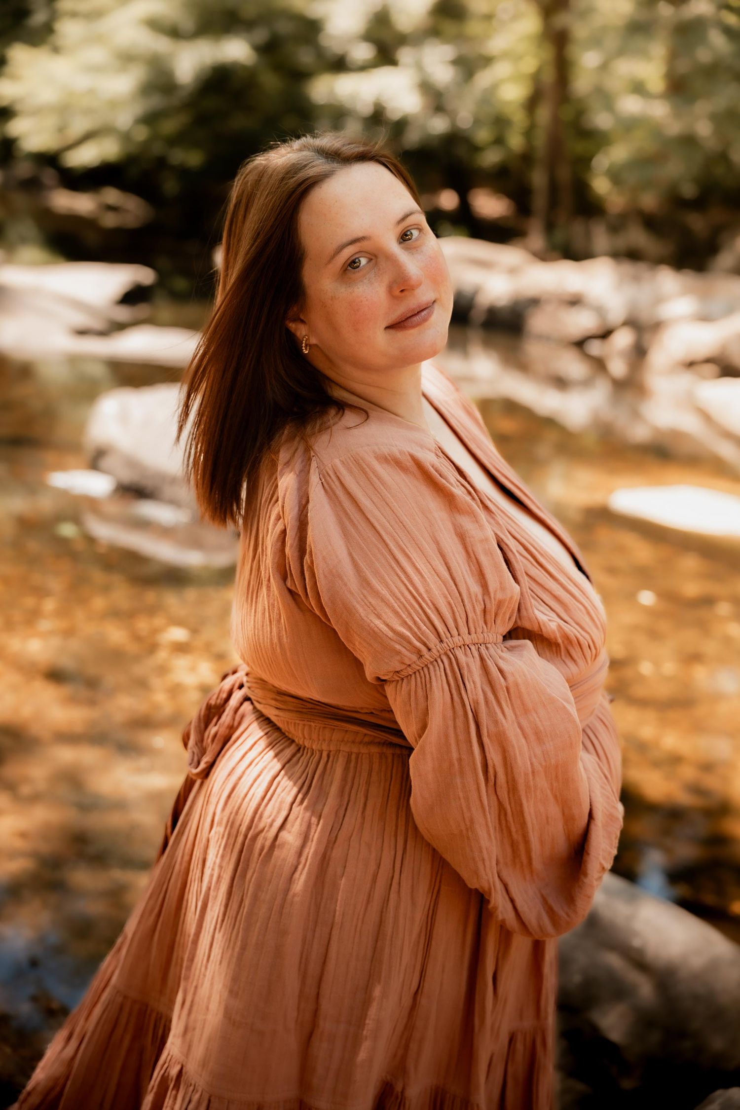 Pregnant woman in a dusty rose dress poses outdoors near a rocky stream surrounded by autumn foliage.