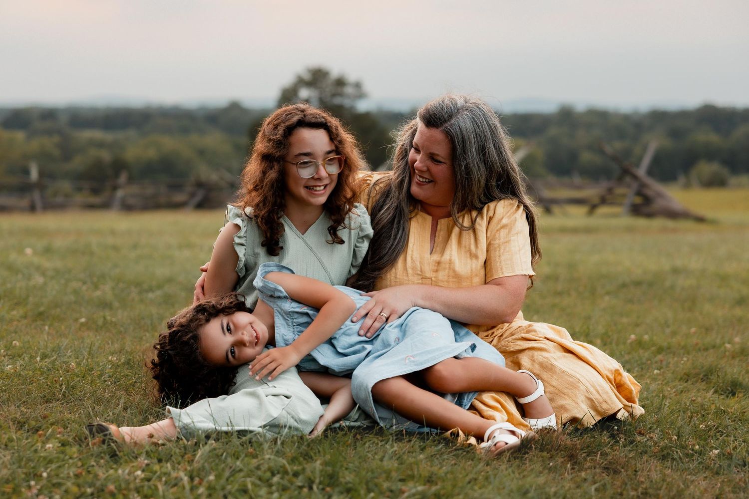 Three women laughing and sitting together on green grass in an outdoor field during a family portrait session.