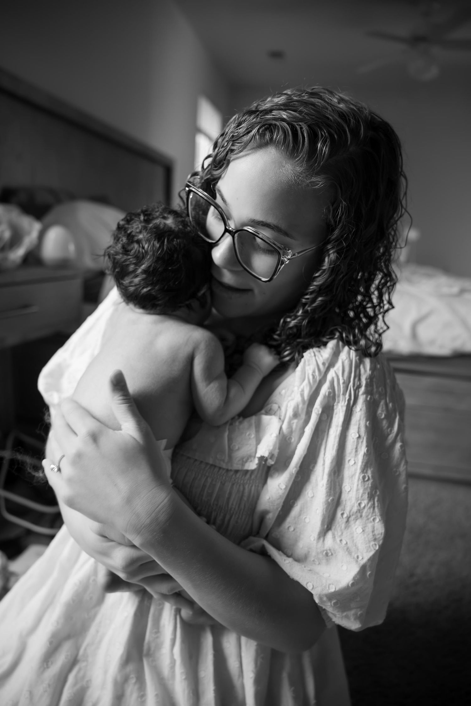 Black and white photo of a curly-haired woman with glasses tenderly holding and kissing a newborn baby indoors.