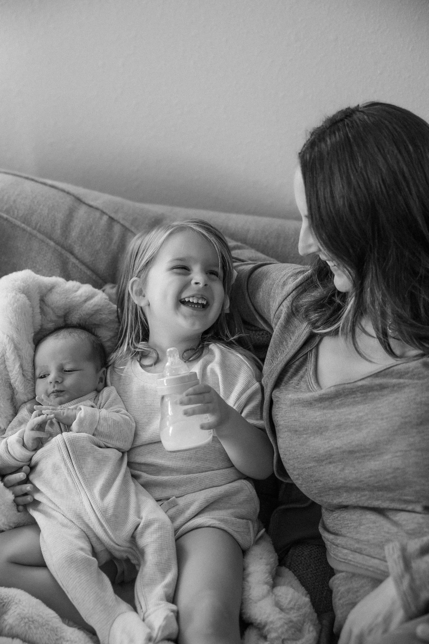 Black and white photo of a laughing toddler holding a bottle sitting with her newborn sibling and mother on a couch.