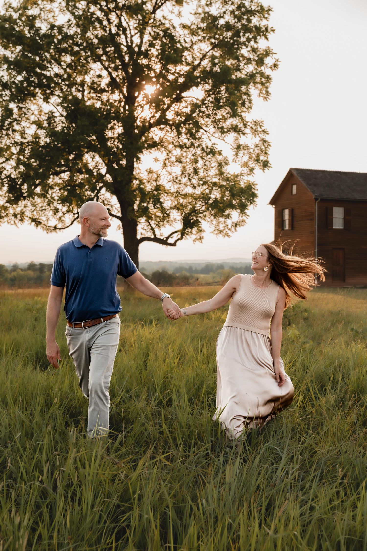 A couple holds hands and walks through tall green grass at sunset near a rustic red barn.