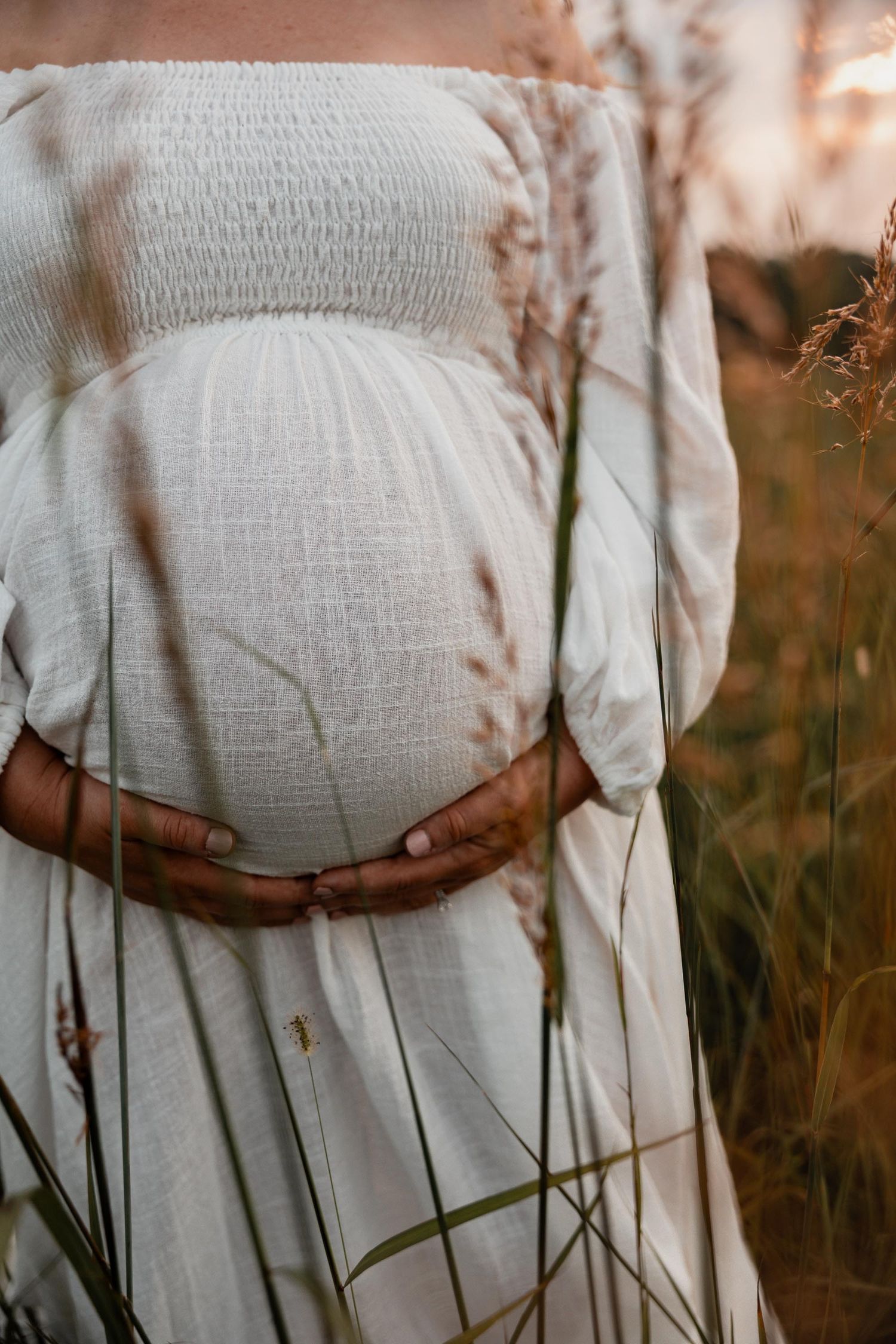 Pregnant woman in white off-shoulder dress cradling baby bump in golden field at sunset.