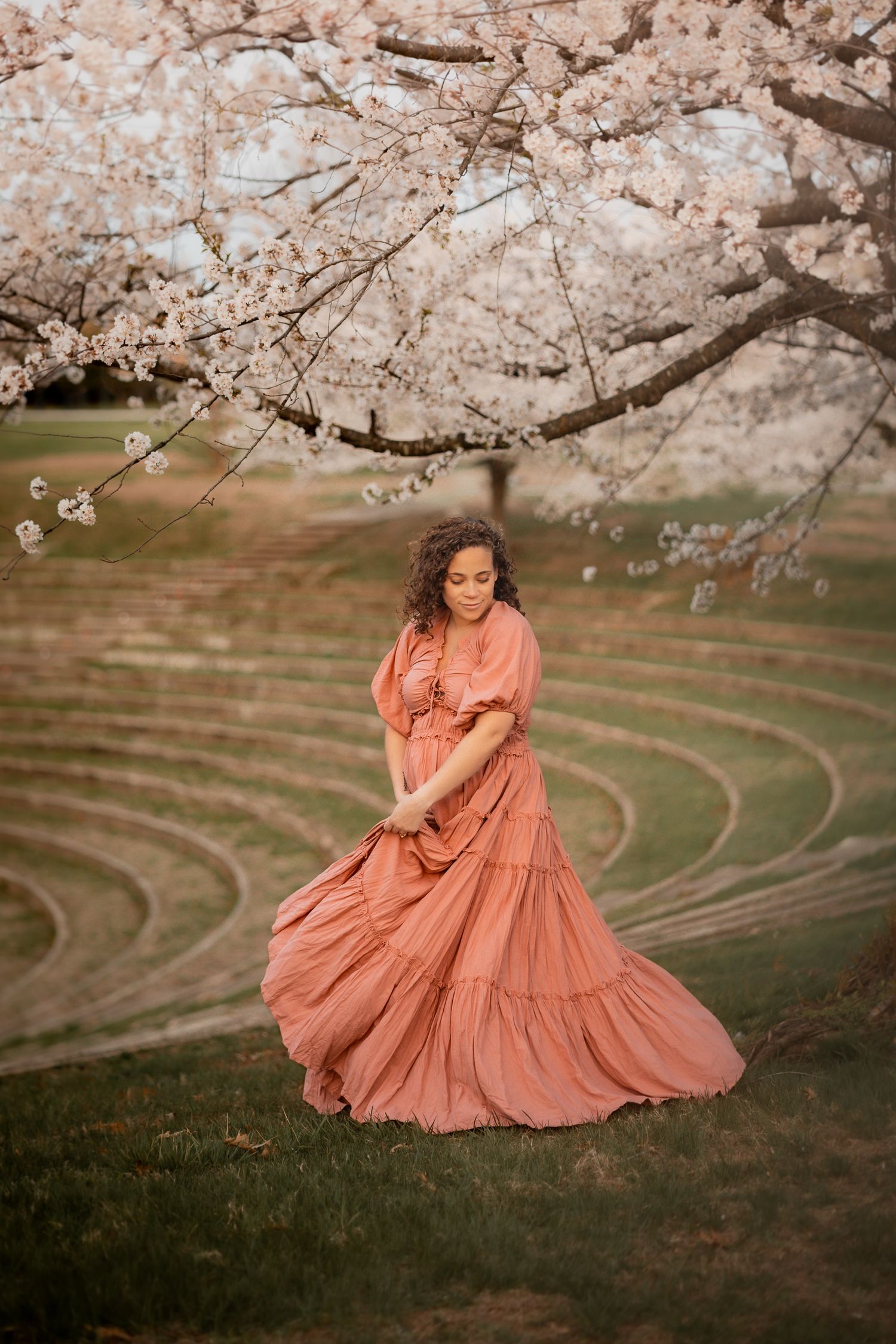 Woman in flowing terracotta dress dancing under blooming cherry blossom tree in spring garden amphitheater.