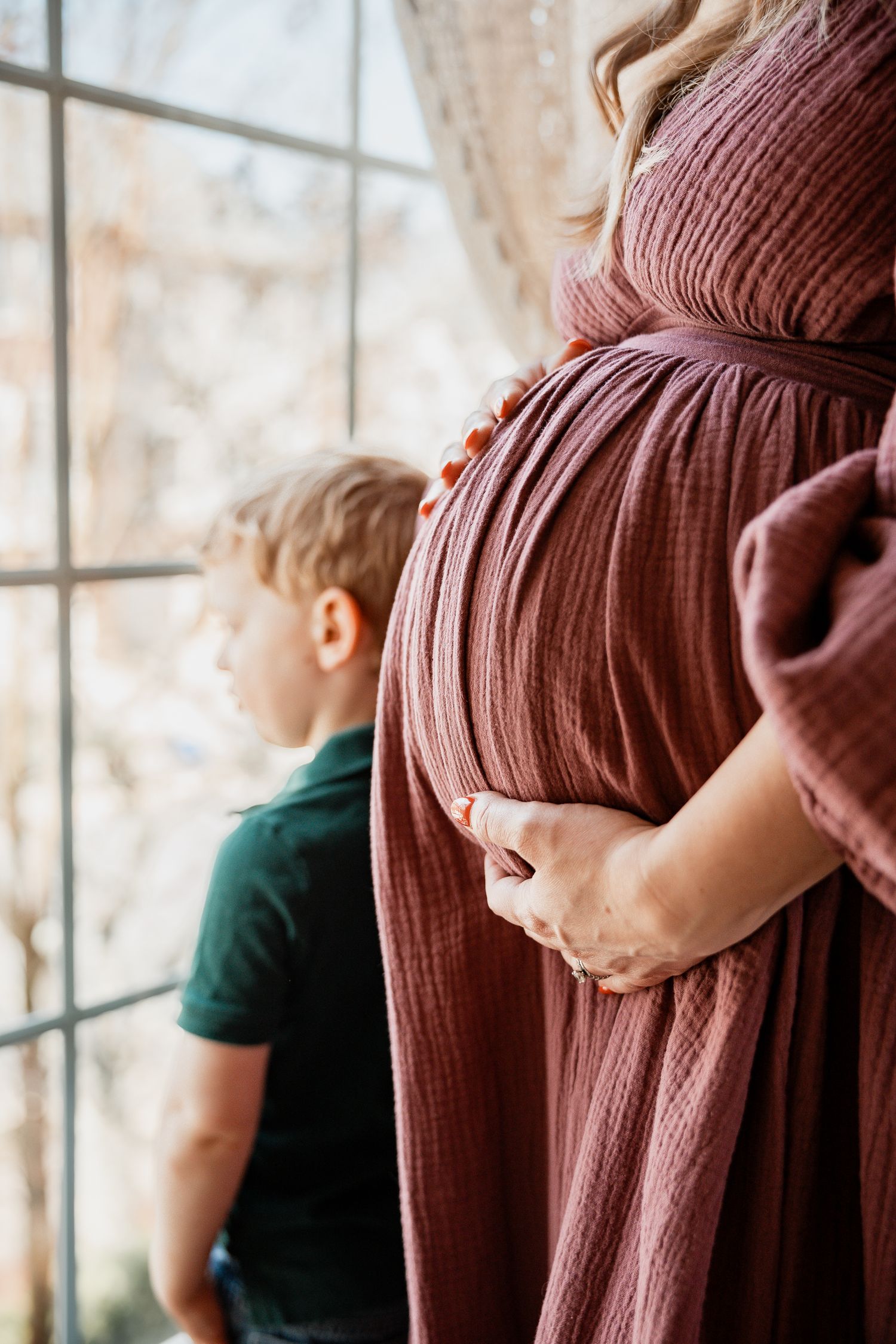 Close-up of pregnant womans baby bump in dusty rose dress with young boy looking out large window.