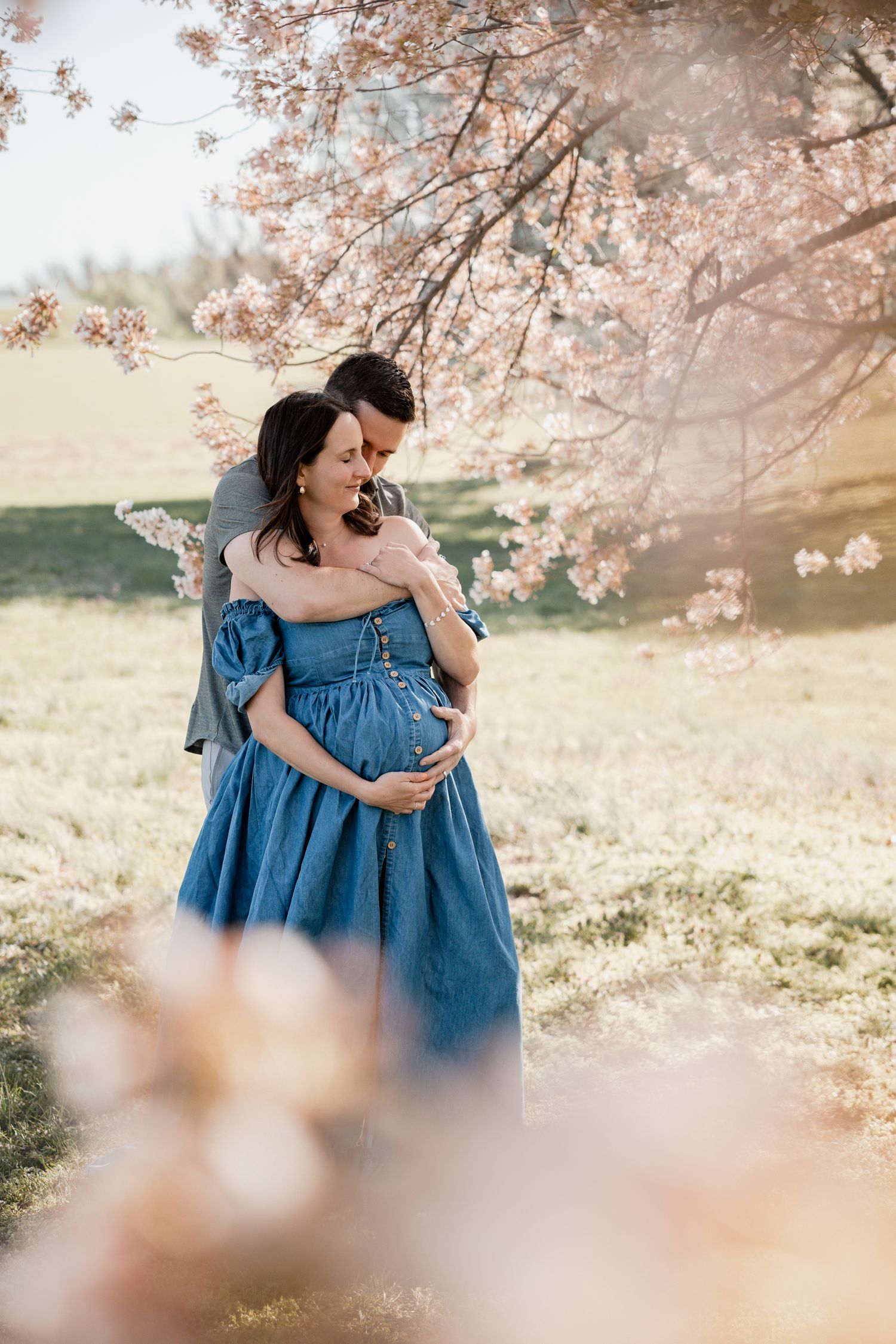 Pregnant couple embracing under cherry blossom tree, woman in blue dress, romantic outdoor maternity photo session.