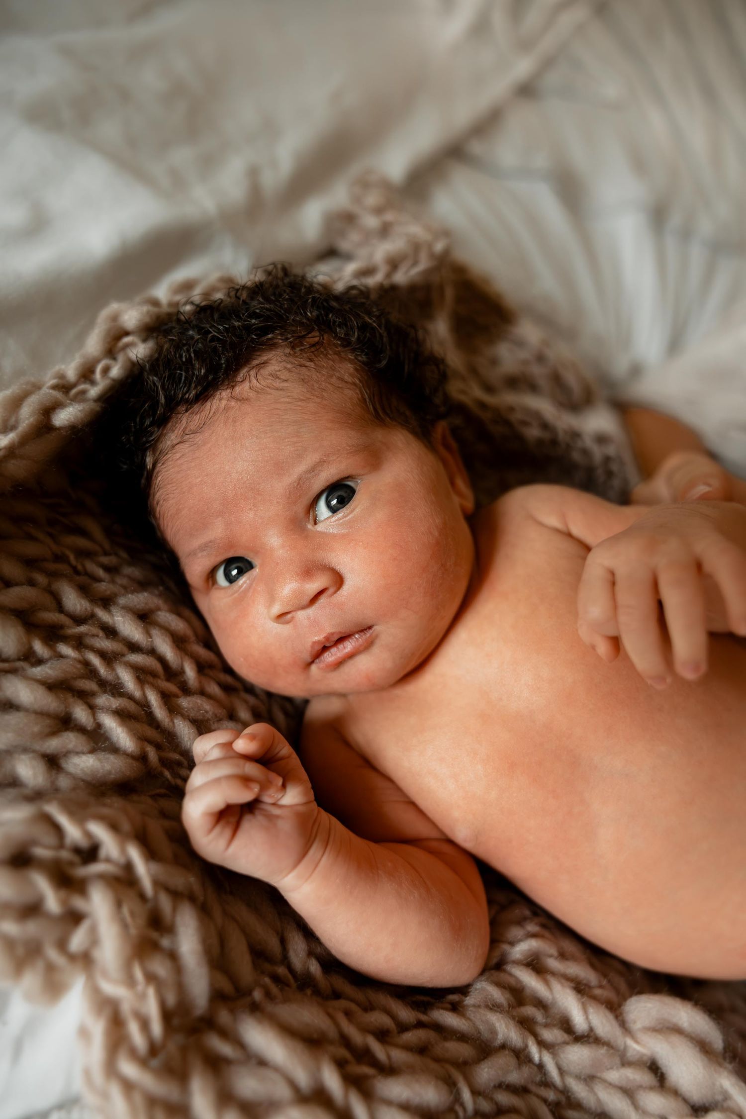 Newborn baby lying on a chunky knit brown blanket, looking up with wide eyes in a soft, natural light setting.