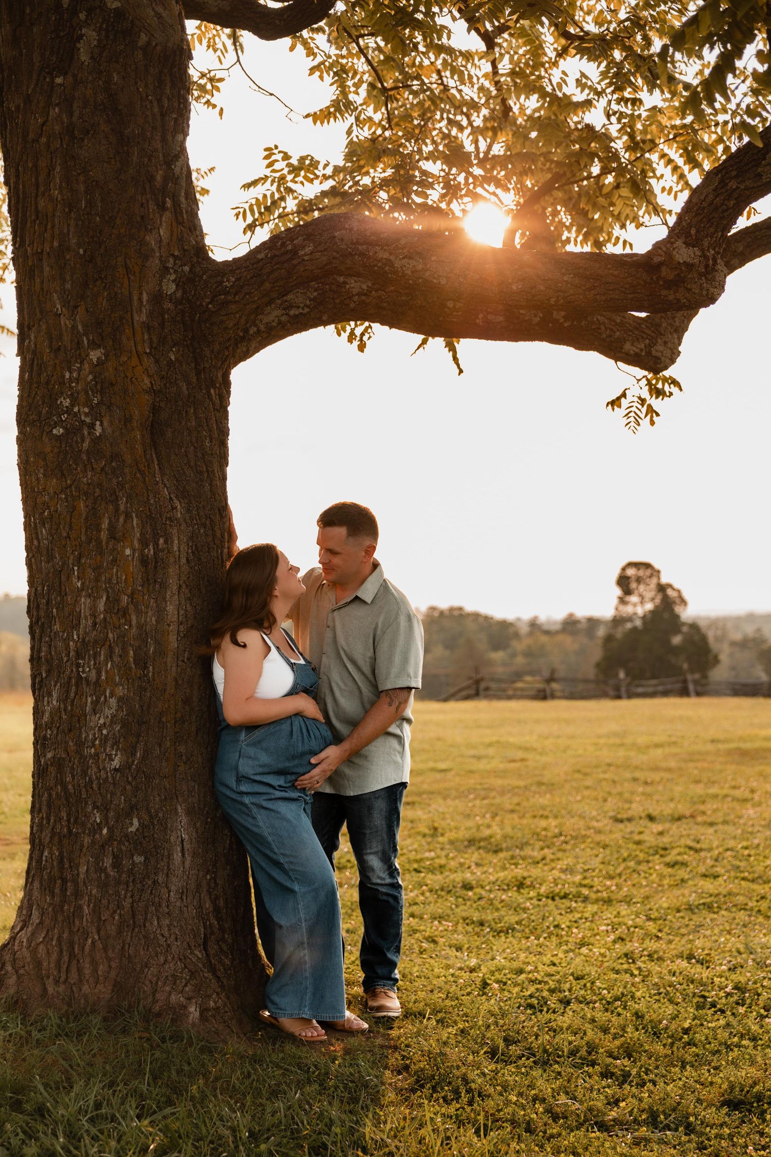 Couple leaning against a large tree at golden hour sunset in a lush green field, sharing a romantic moment.