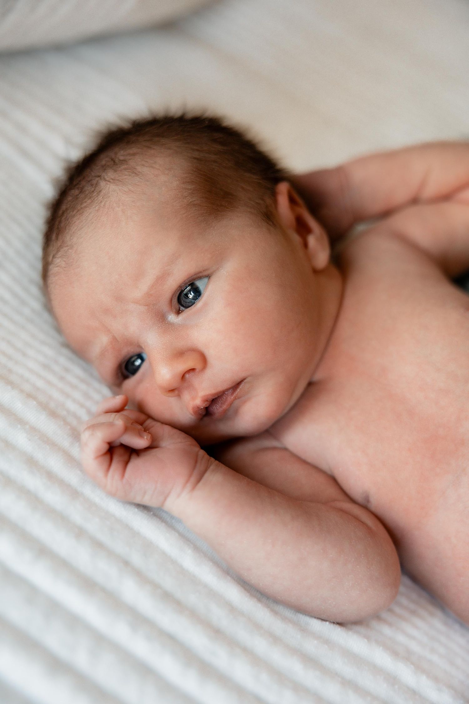 Awake newborn baby with dark hair lying on a white blanket, gazing curiously with bright blue eyes.