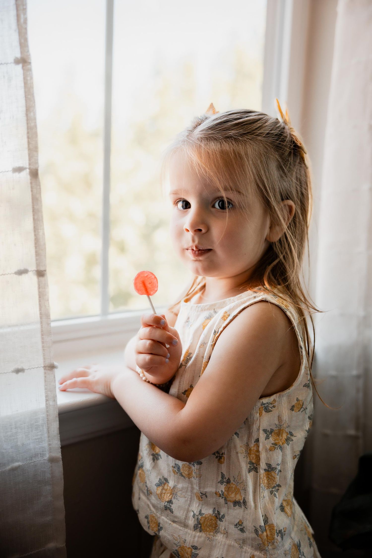Cute toddler girl holding a lollipop while standing by a sunny window.