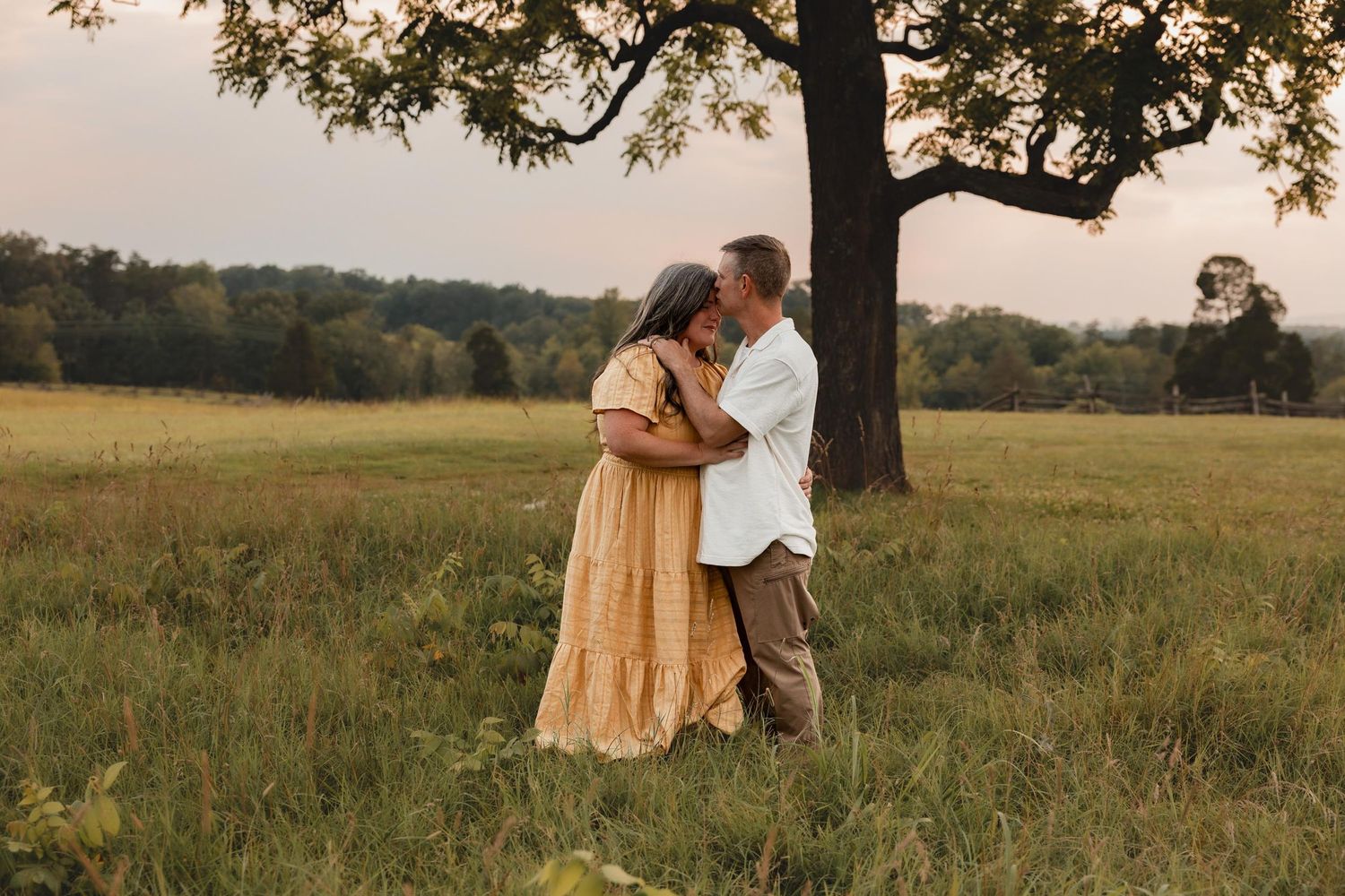 Couple embracing and kissing in a lush green meadow under a large tree at golden hour.