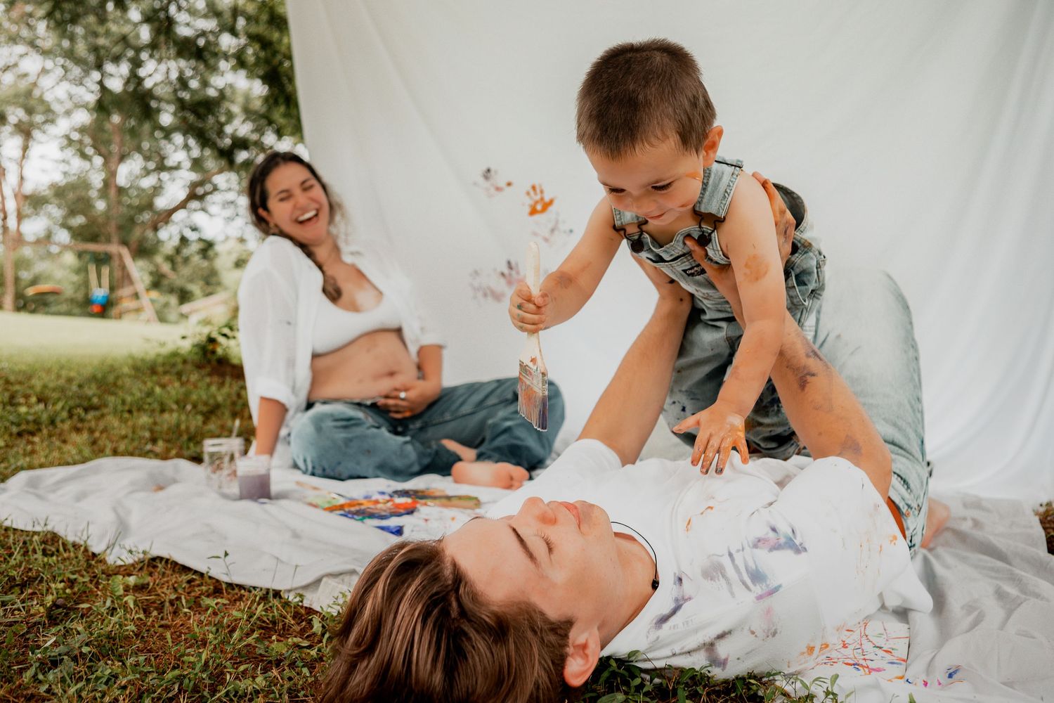 Family lounging together with baby on white blanket in outdoor garden setting during summer photo session.