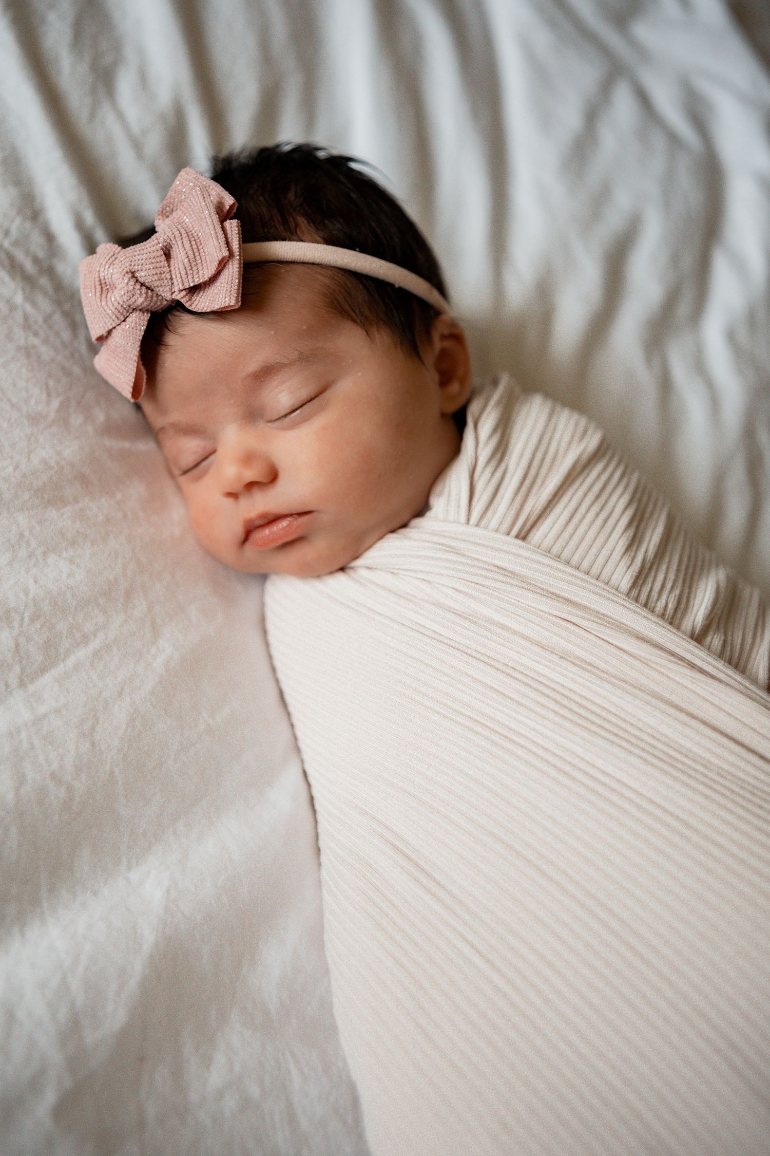 Peaceful sleeping newborn baby wearing white clothing and pink bow headband on white bedding.