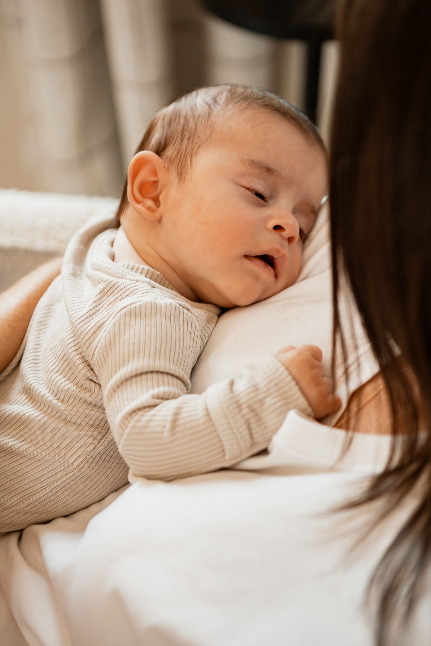 A peaceful newborn baby sleeps soundly on white bedding while wearing a cozy cream-colored sweater.