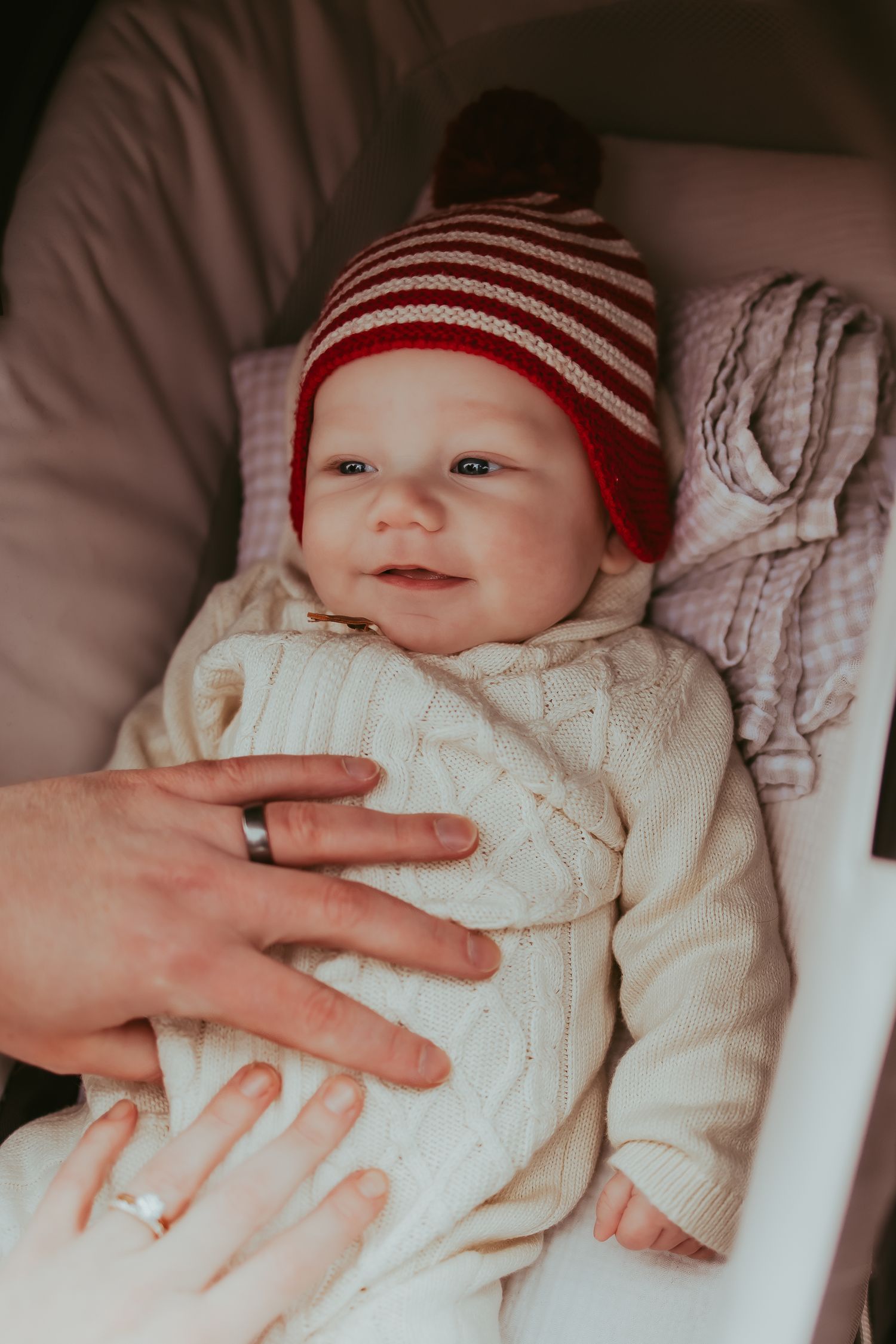 A baby wearing a red and white striped hat lies in a white knit sweater with an adult's hand resting gently on them.