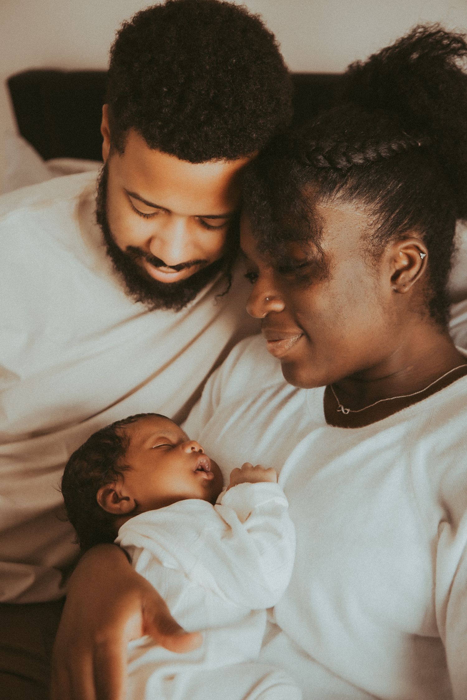 A tender family moment shows parents in white shirts smiling lovingly at their sleeping baby in a warm-toned photograph.