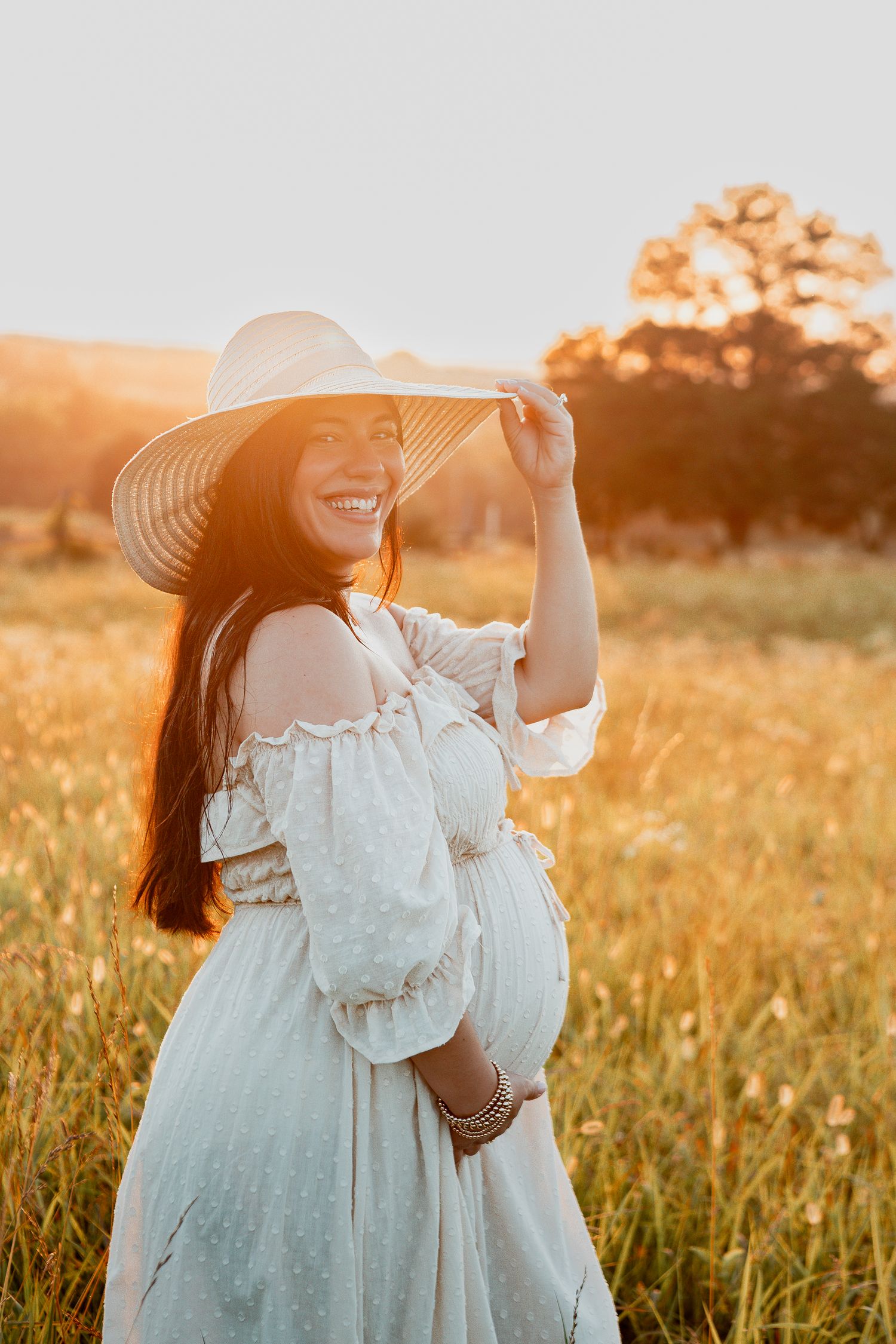 Woman in white dress and sun hat poses in golden field during sunset maternity photoshoot.