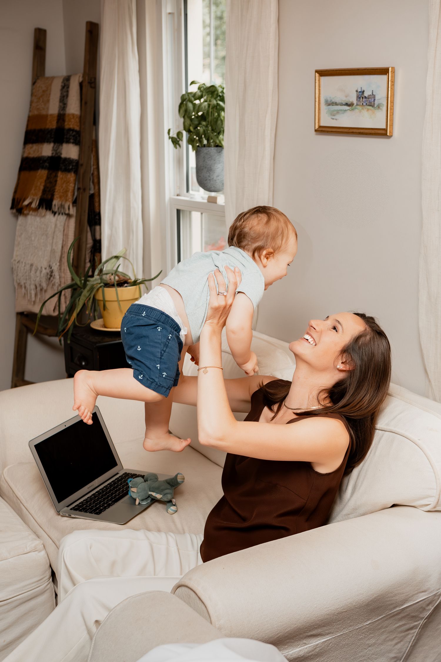 A parent and baby share playful moments while working from home on a white couch with laptop nearby.