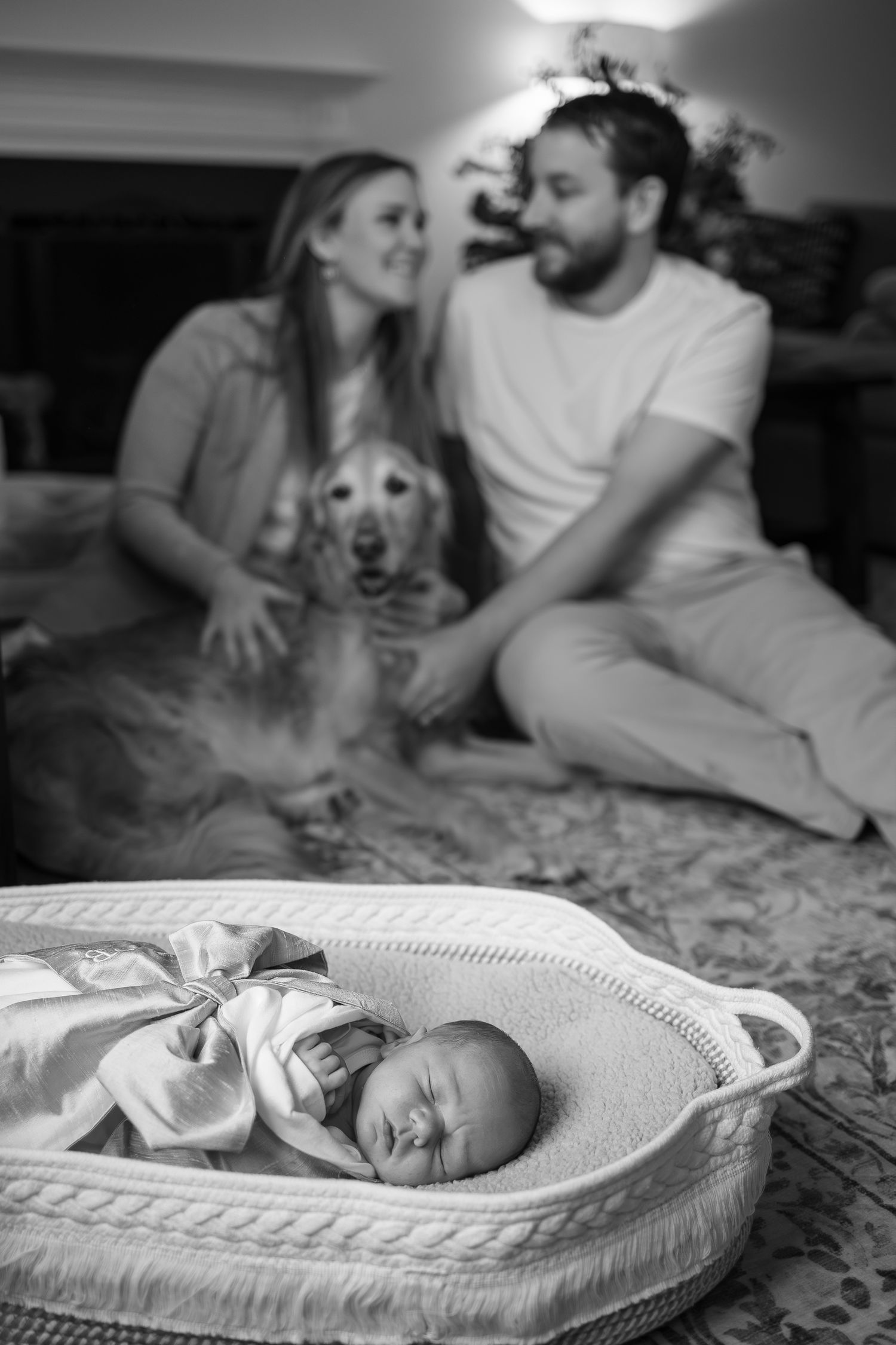 Black and white photo of newborn in basket with couple and dog in background.