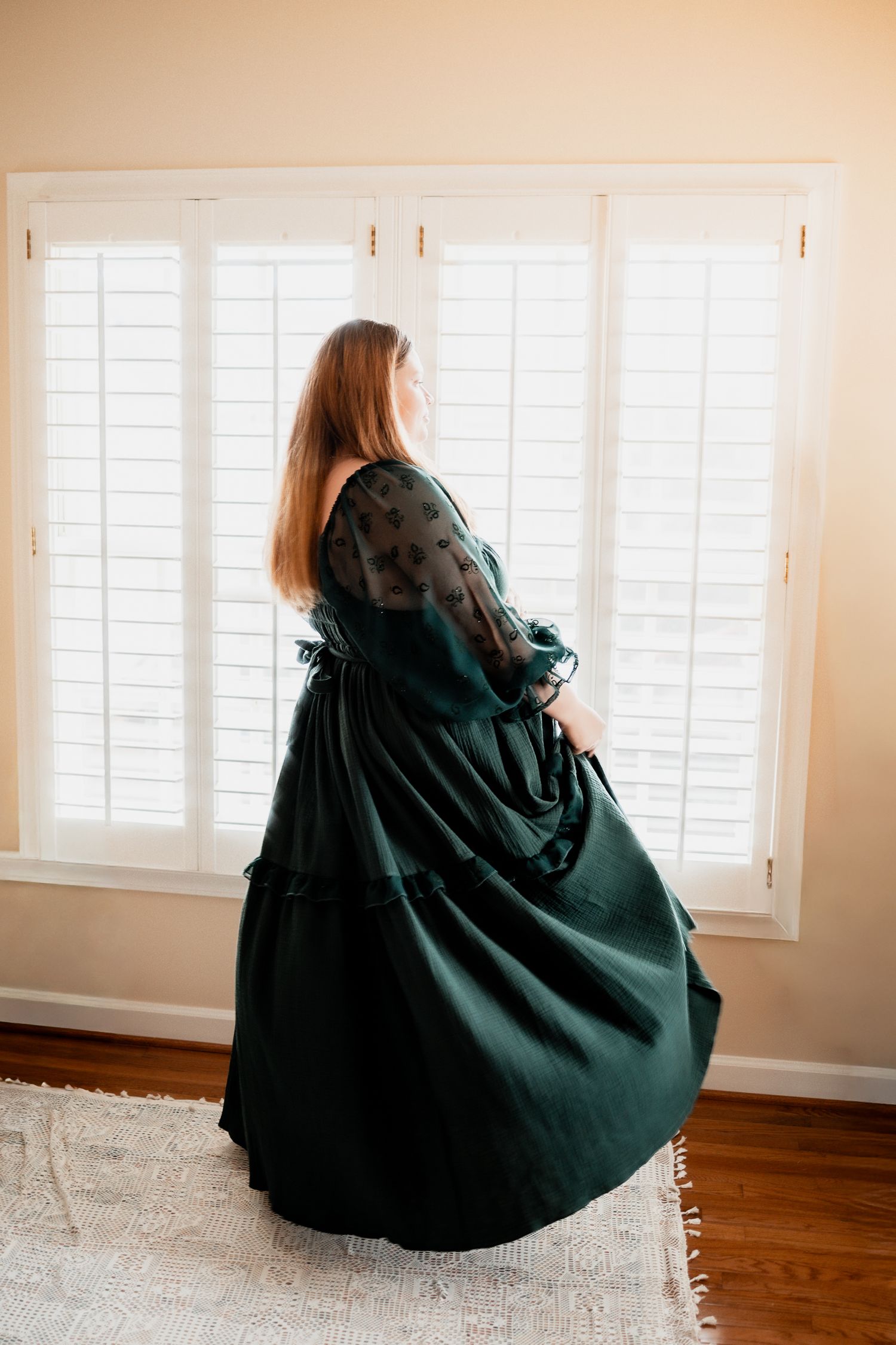 Woman in elegant dark green Victorian ball gown standing by white plantation shutters in a sunlit room. Maternity DMV