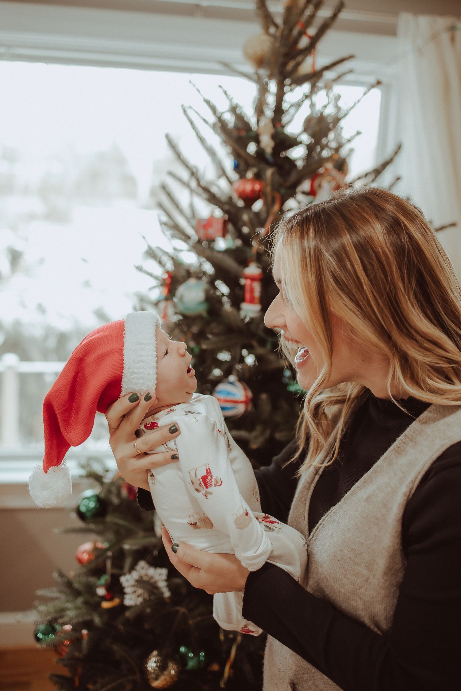 Two friends laugh and share hot beverages in front of a decorated Christmas tree during the holiday season.