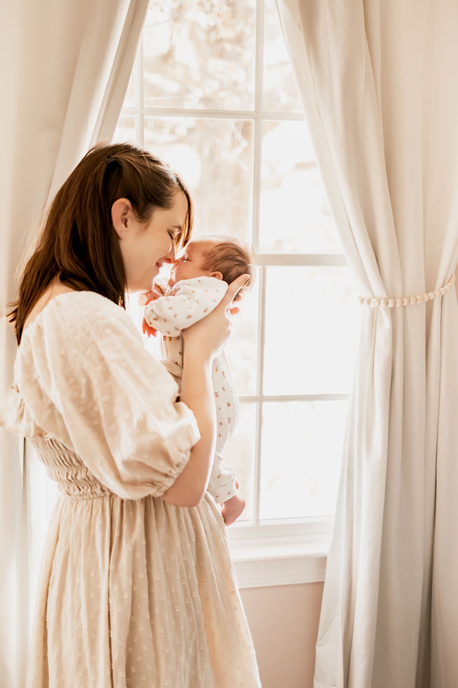 A tender moment by a bright window as a woman in a vintage cream dress holds a newborn baby in her arms.