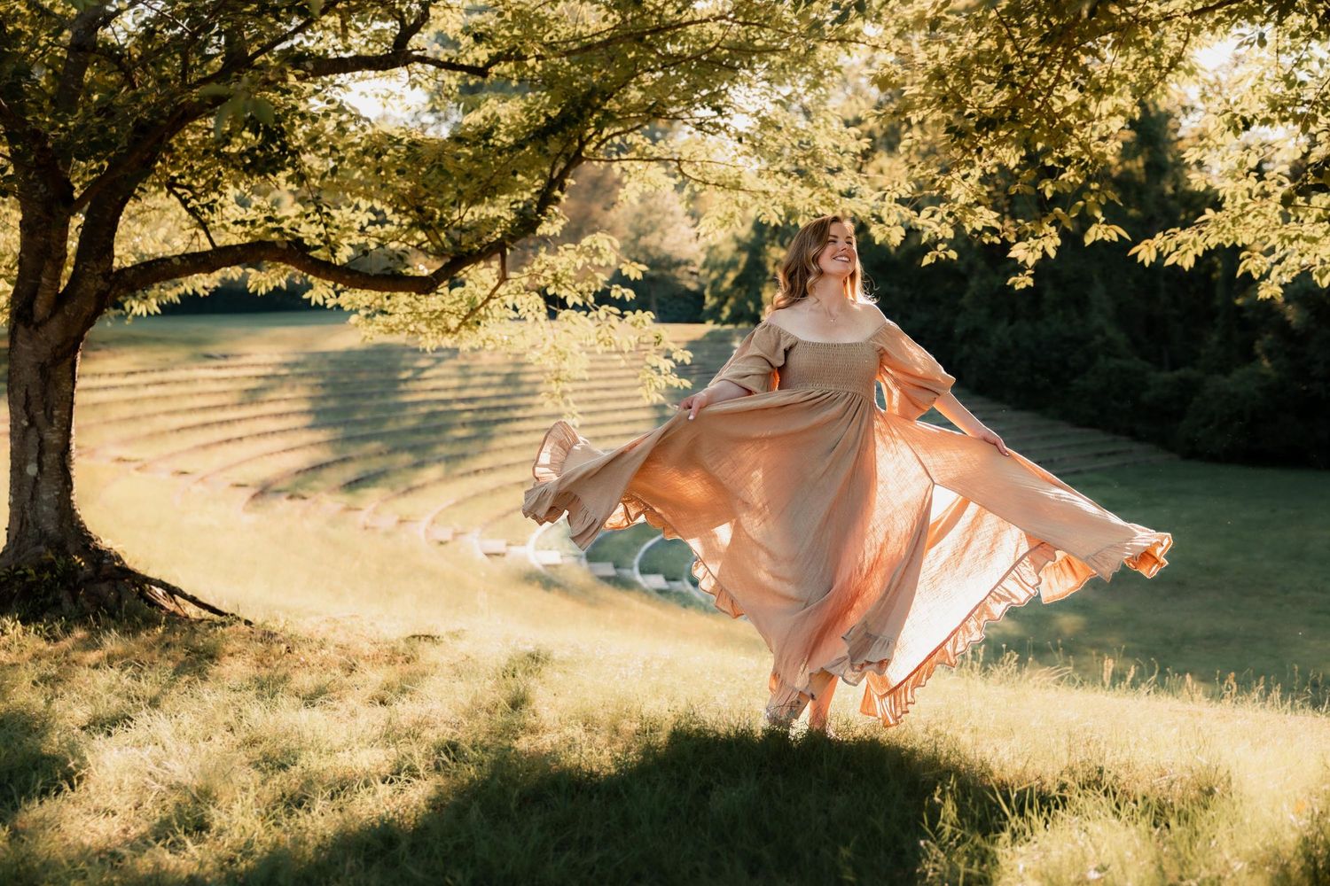 Woman in flowing peach gown twirling under sunlit trees in a lush green garden.