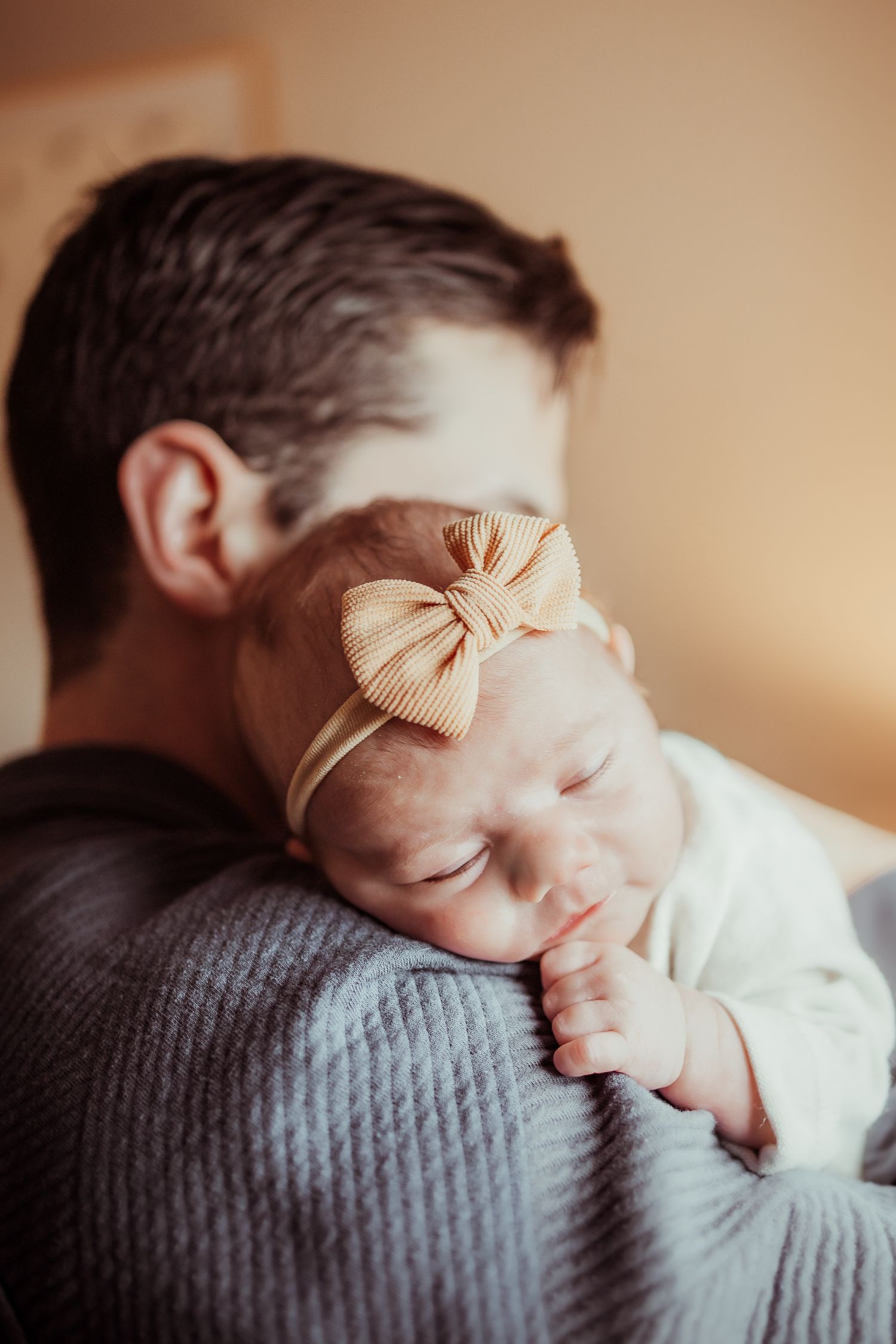 A sleeping newborn wearing a peach bow headband rests peacefully on a gray textured blanket.