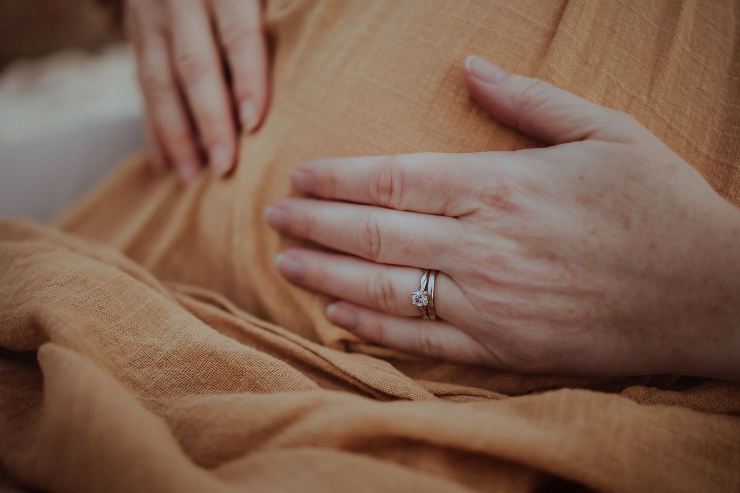 Close up of hands with wedding ring resting on soft peach colored fabric in gentle lighting.