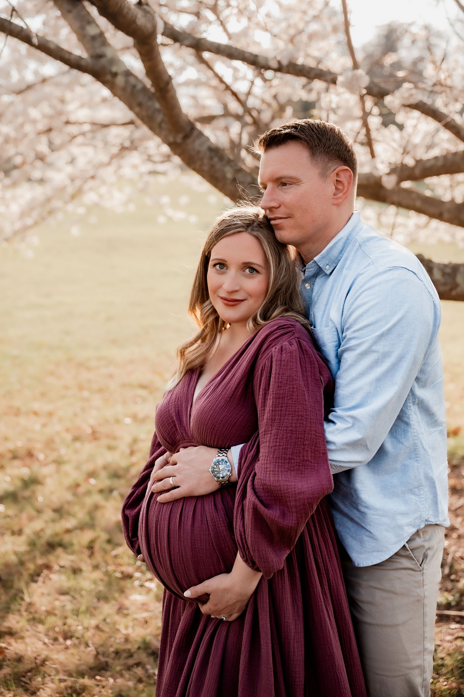 Couple sharing a tender maternity photo outdoors under blooming cherry blossom trees, woman in burgundy dress cradling baby bump.