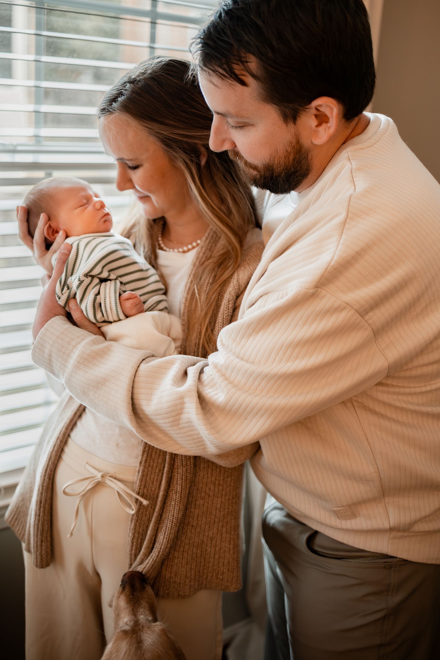 Couple in warm sweaters holding their newborn baby near window with natural light.