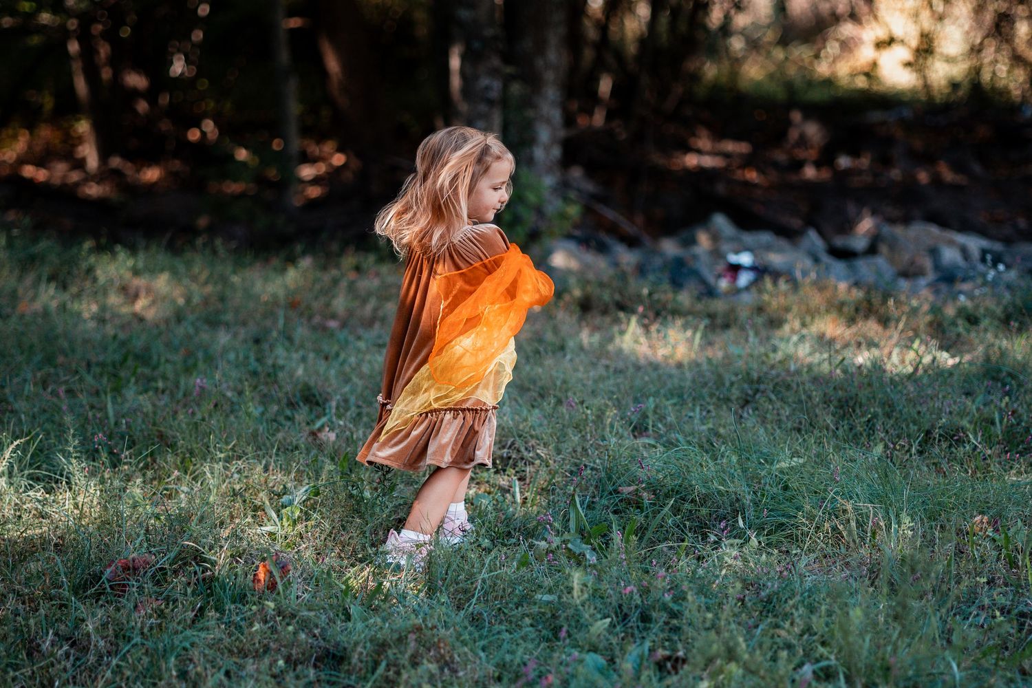 A young person in a pink dress and orange boots plays joyfully in a grassy outdoor setting at sunset.