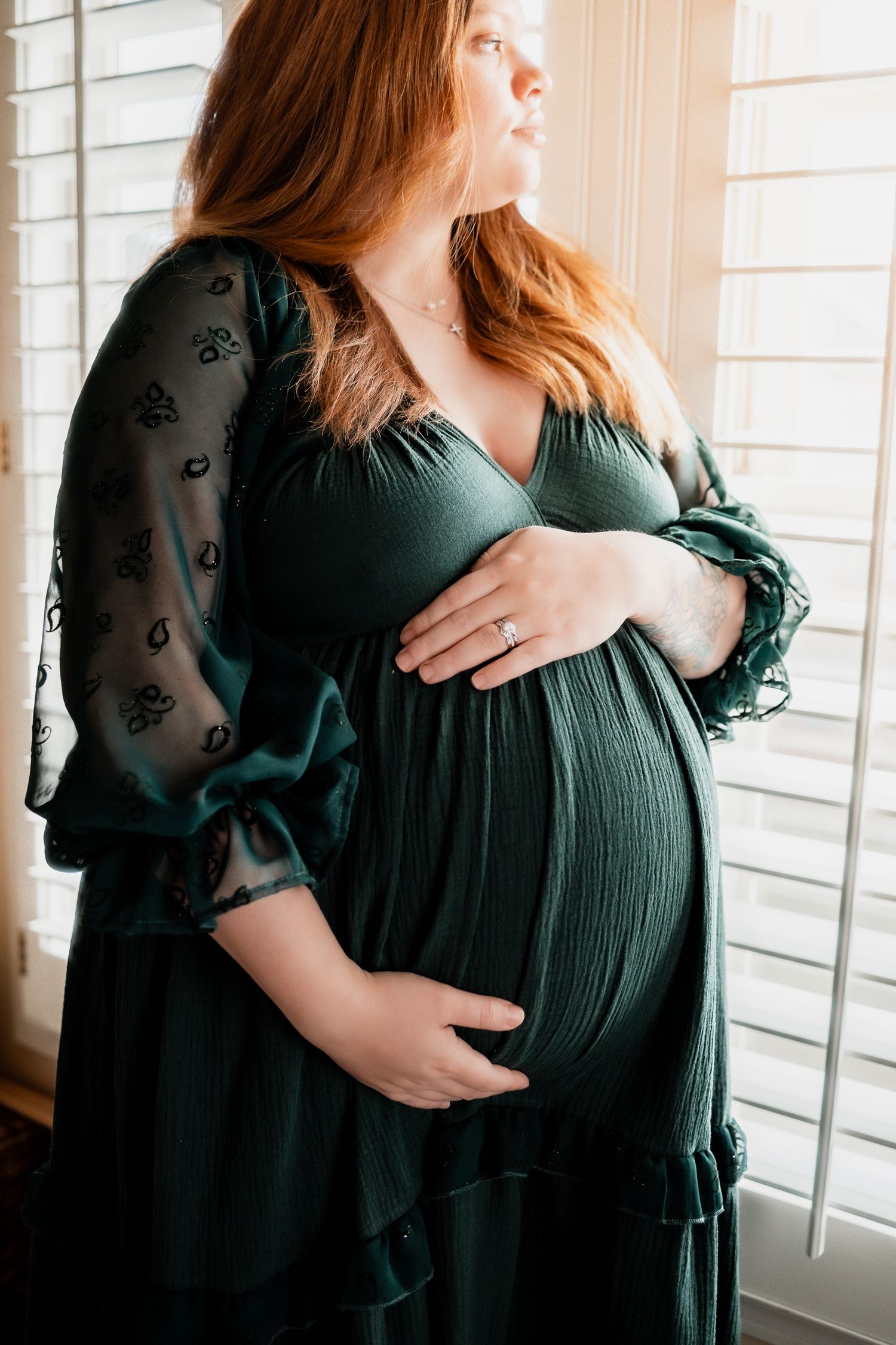 Pregnant woman in green dress cradling baby bump by window, maternity photography with natural light. Maternity DMV