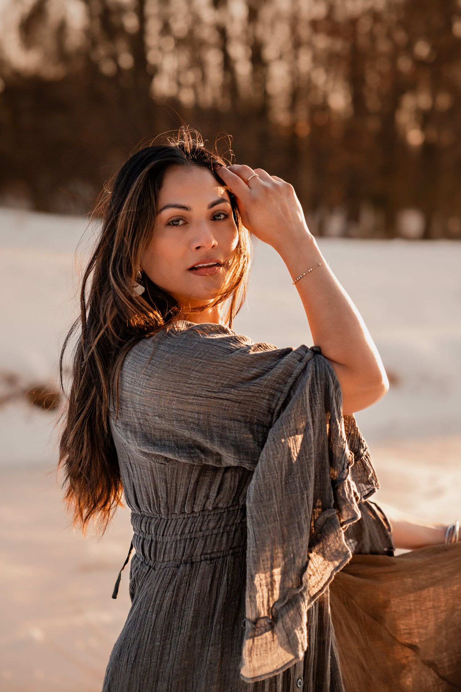 A person in a gray dress poses playfully in a snowy winter landscape during golden hour lighting.