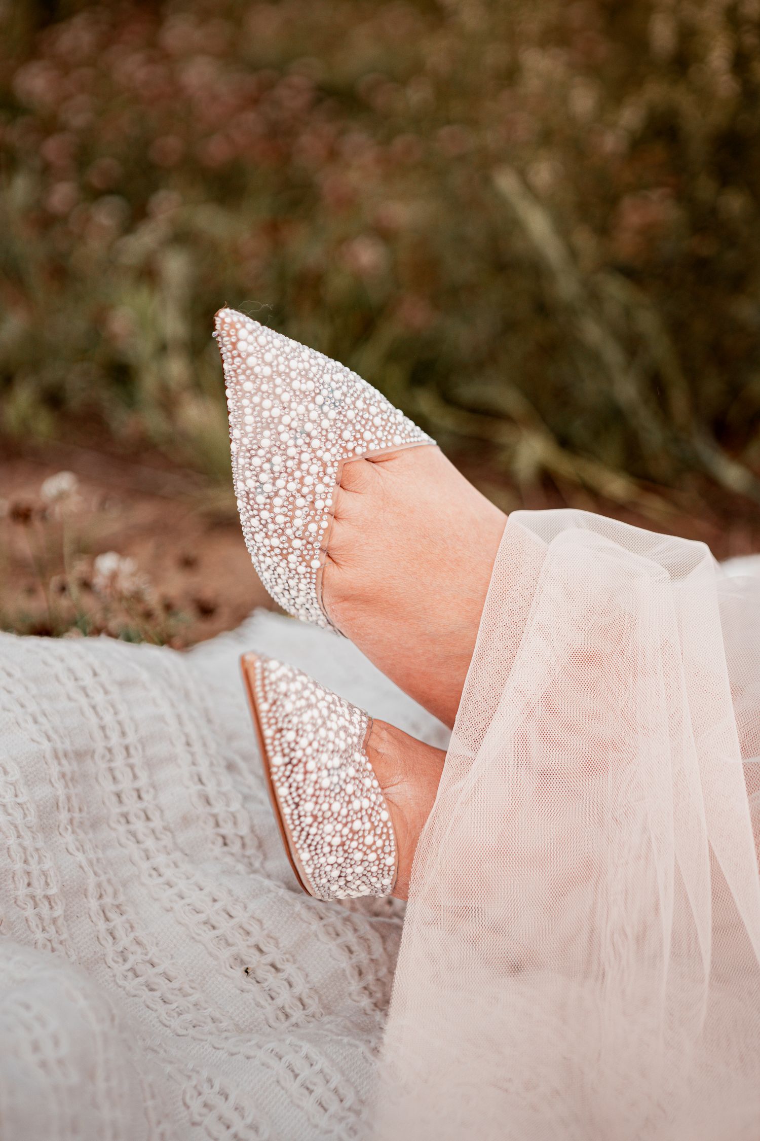 A sparkling silver glitter bridal shoe with sheer tulle fabric detail rests on a textured white lace surface.