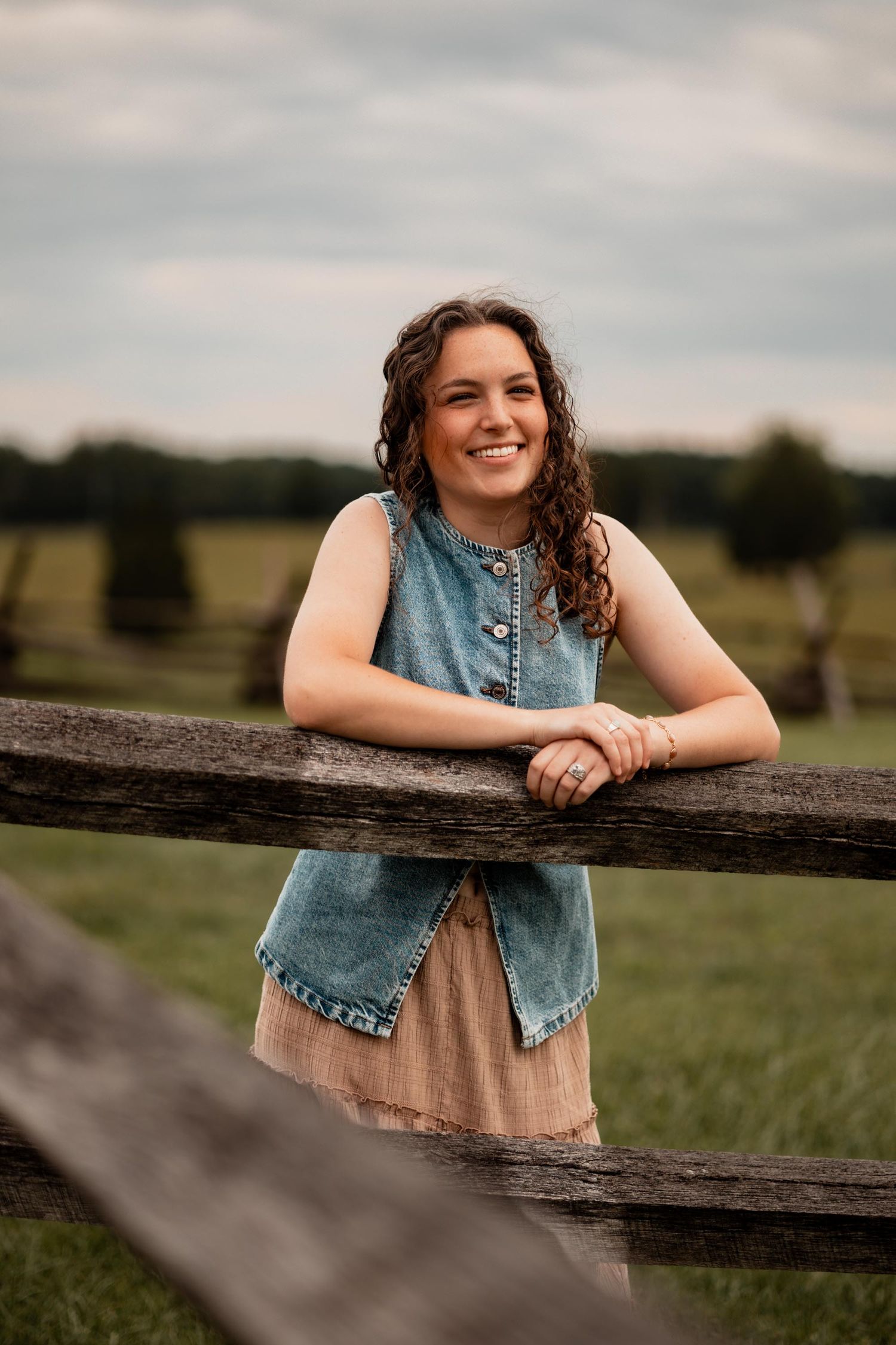 Smiling young woman in denim vest leaning on rustic wooden fence in green countryside field.