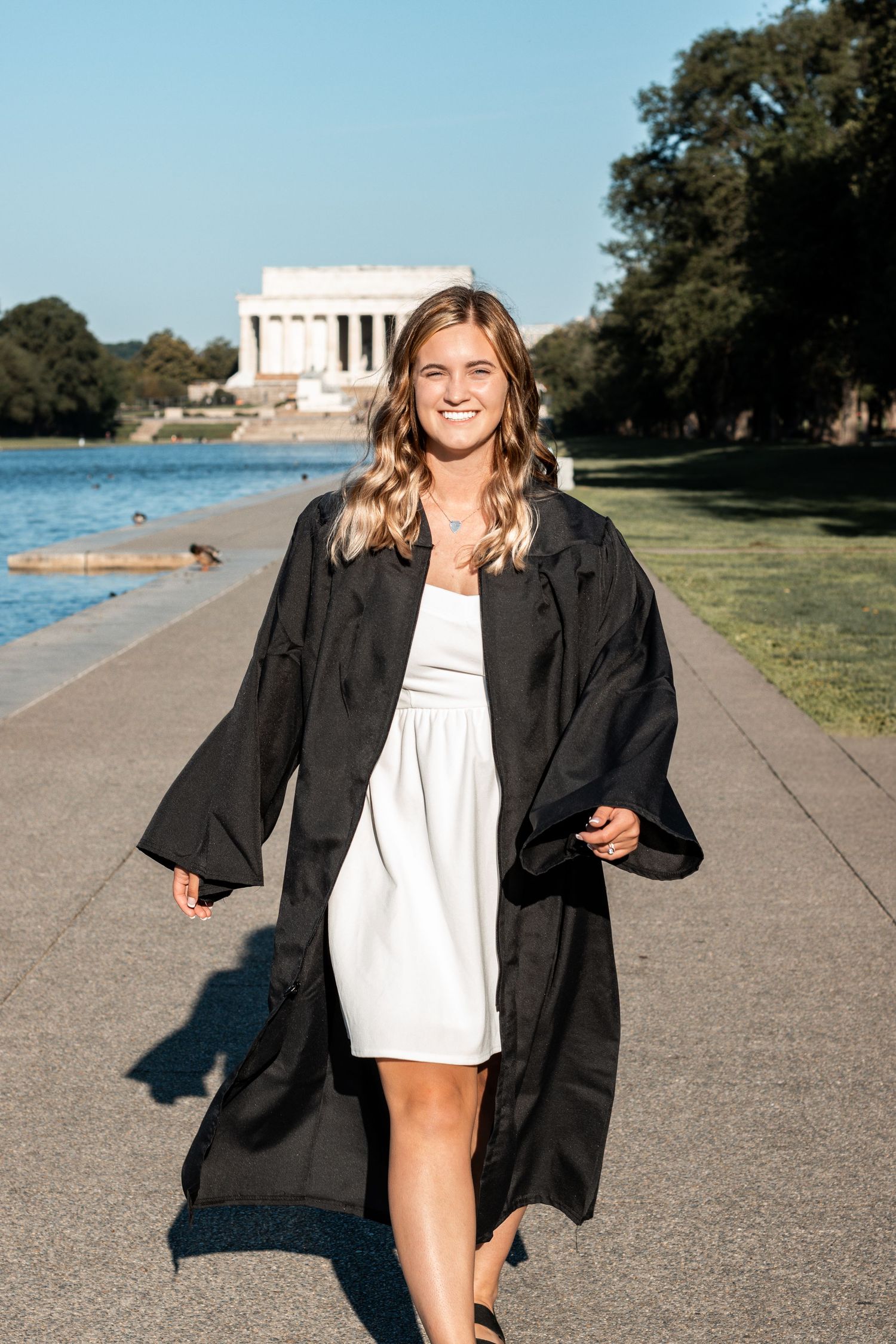 Graduate in black gown walks along reflecting pool path with Lincoln Memorial in background.