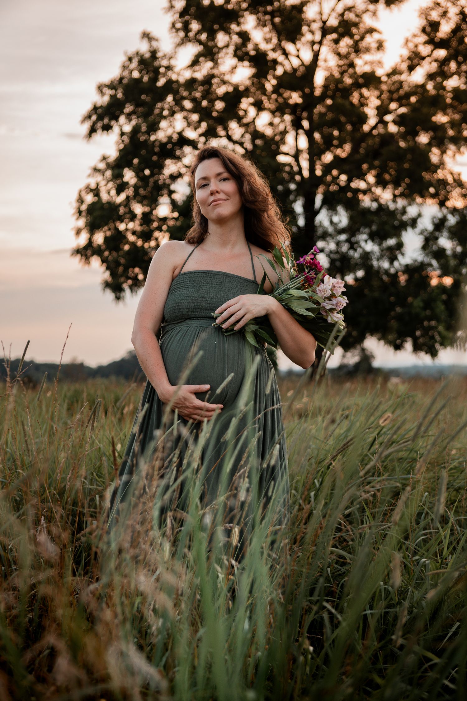 A maternity portrait in a field at sunset features an expectant mother in a sage green dress holding wildflowers.