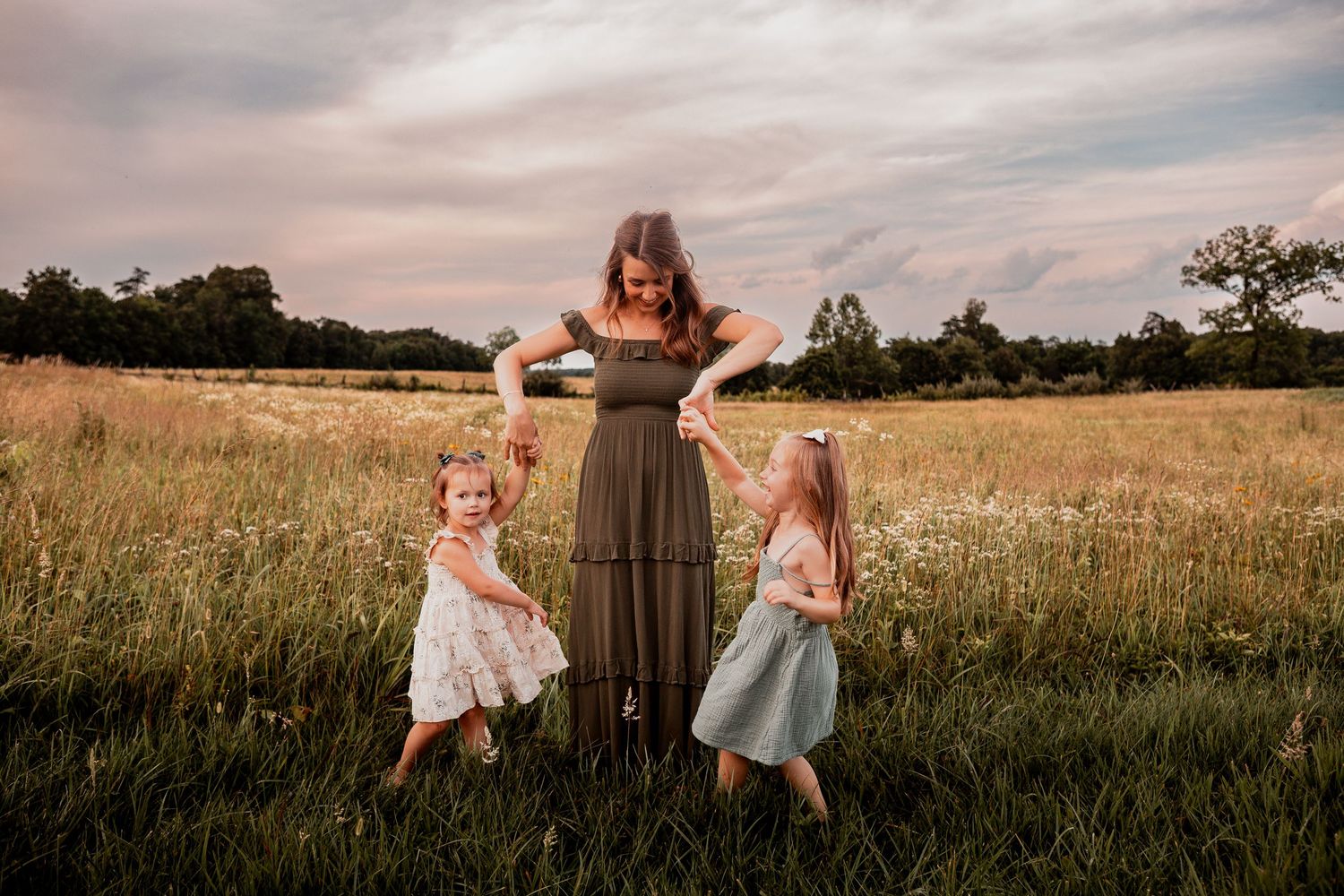 Children in sundresses play and run through a golden field at sunset with dramatic cloudy skies overhead.