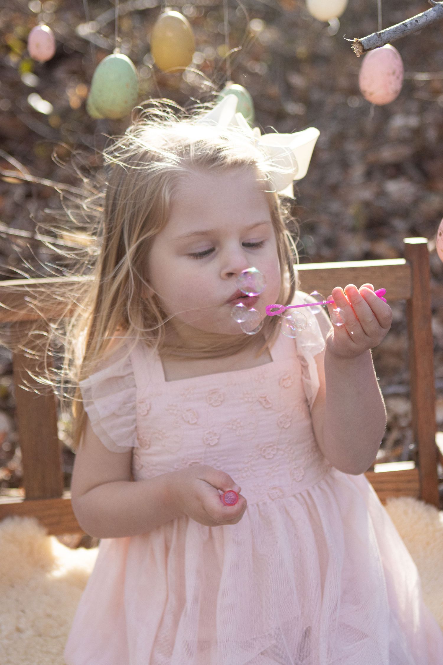 Young child in pink dress blowing on dandelions with Easter eggs hanging in background.