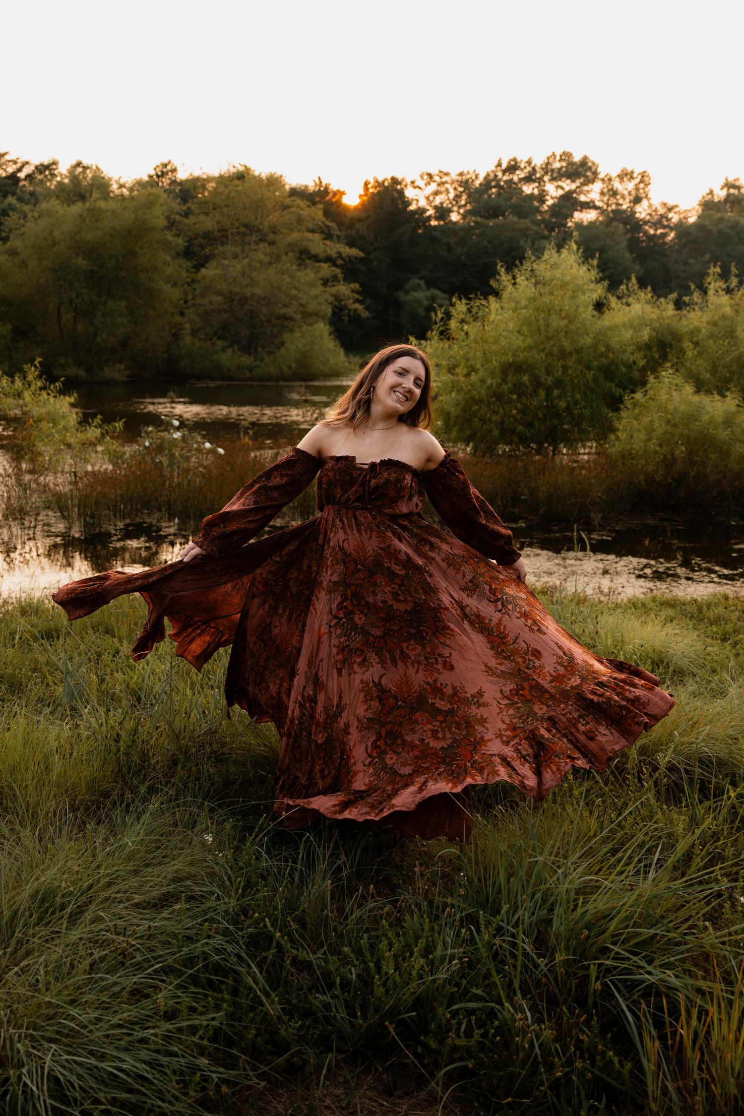 Woman in flowing rust floral off-shoulder dress twirling in golden hour light near a serene pond.