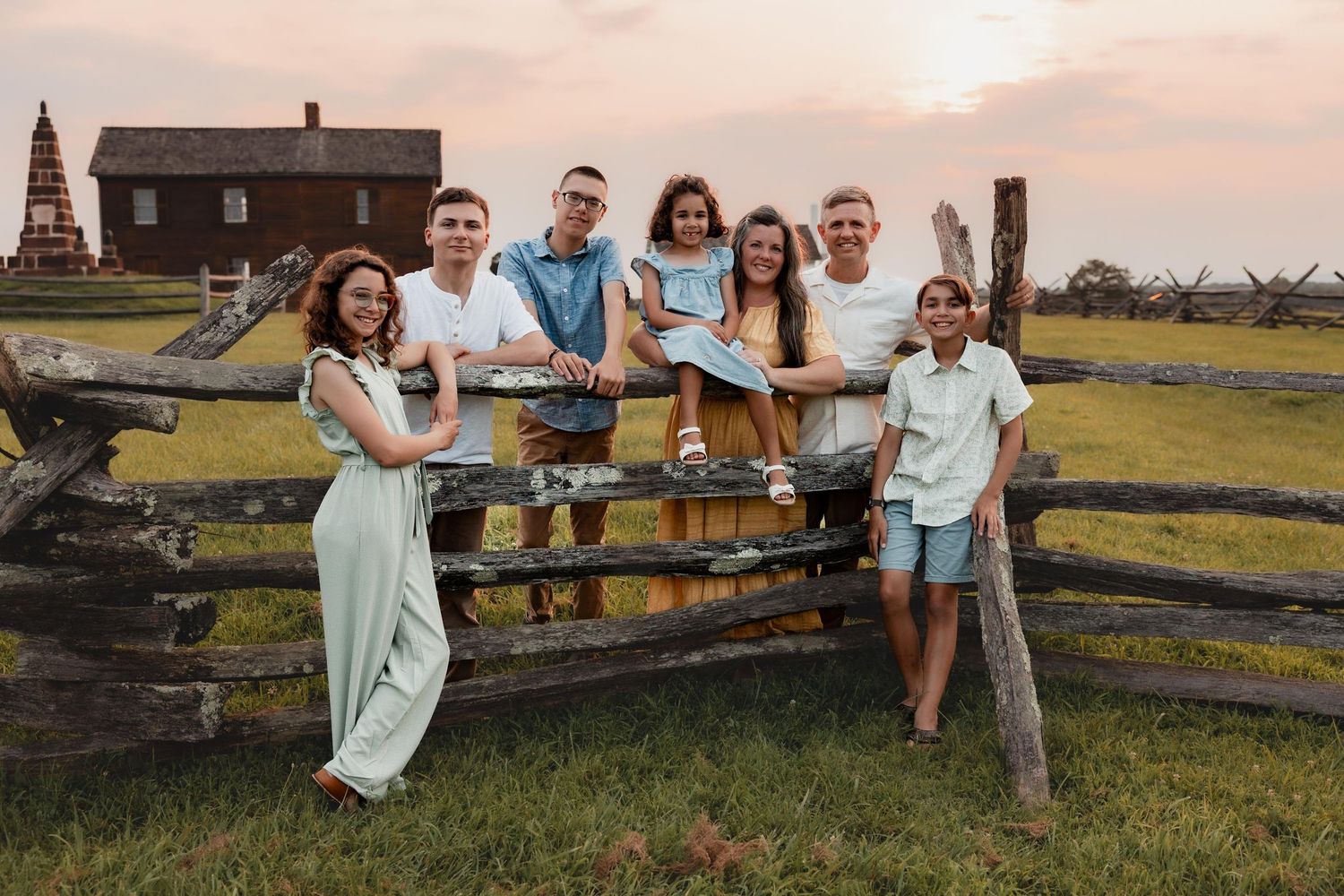 Family of seven posing by a rustic wooden fence at golden hour with historic buildings in the background.