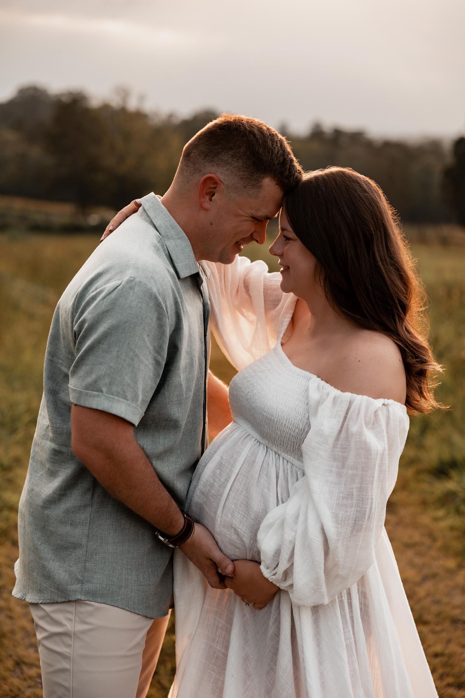 Couple sharing a tender forehead touch moment in a field at golden hour, woman in white off-shoulder maternity dress.