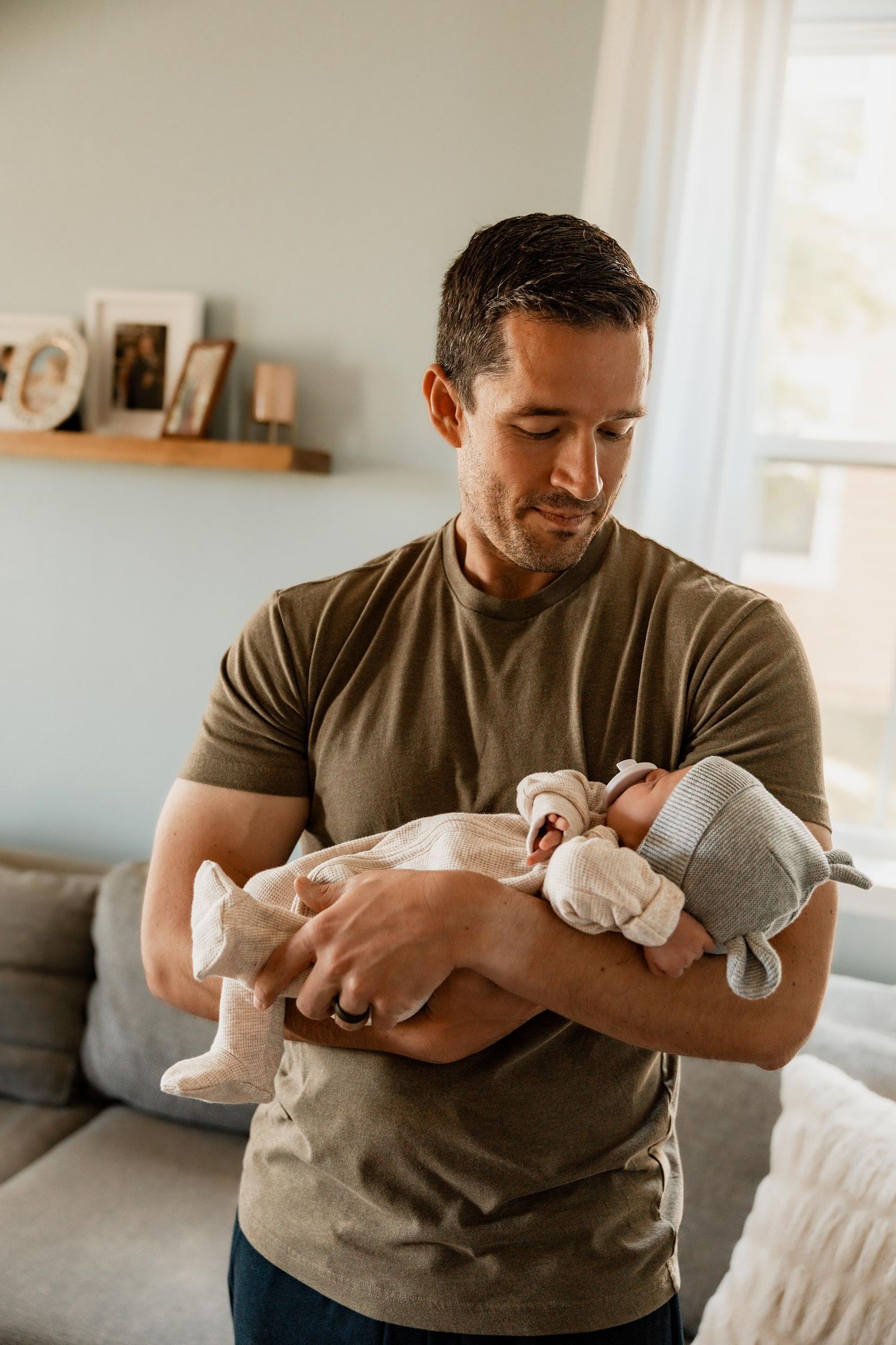 Father in olive green t-shirt gently cradling sleeping newborn baby in his arms in a bright living room.