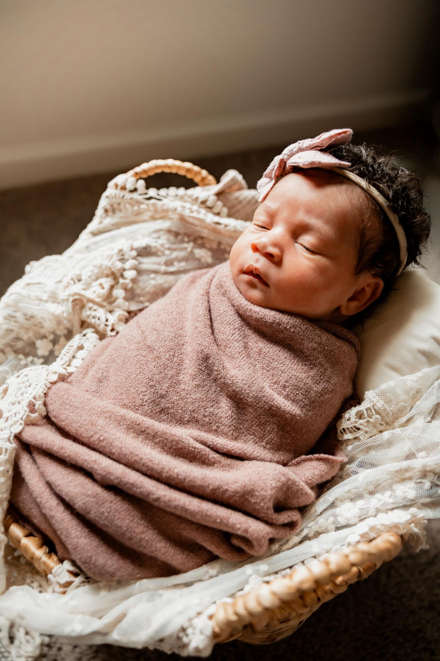 Sleeping newborn baby girl swaddled in pink wrap with bow headband resting in a wicker bassinet.