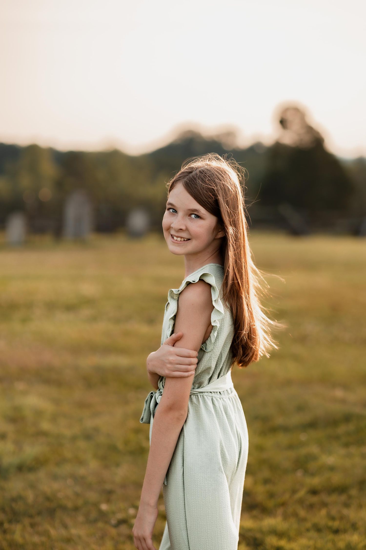 A smiling young girl with long brown hair wearing a mint green jumpsuit poses in a sunlit green field at golden hour.