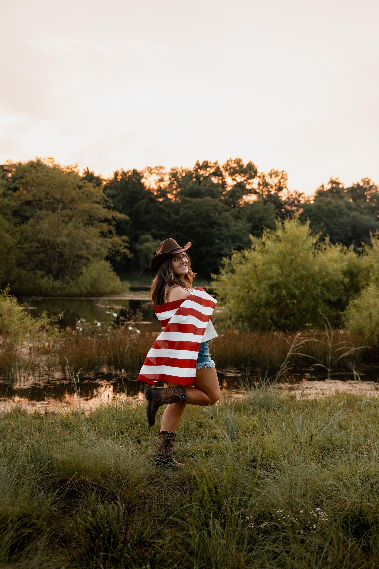 Woman in cowboy hat wrapped in American flag standing in green field at golden hour sunset.