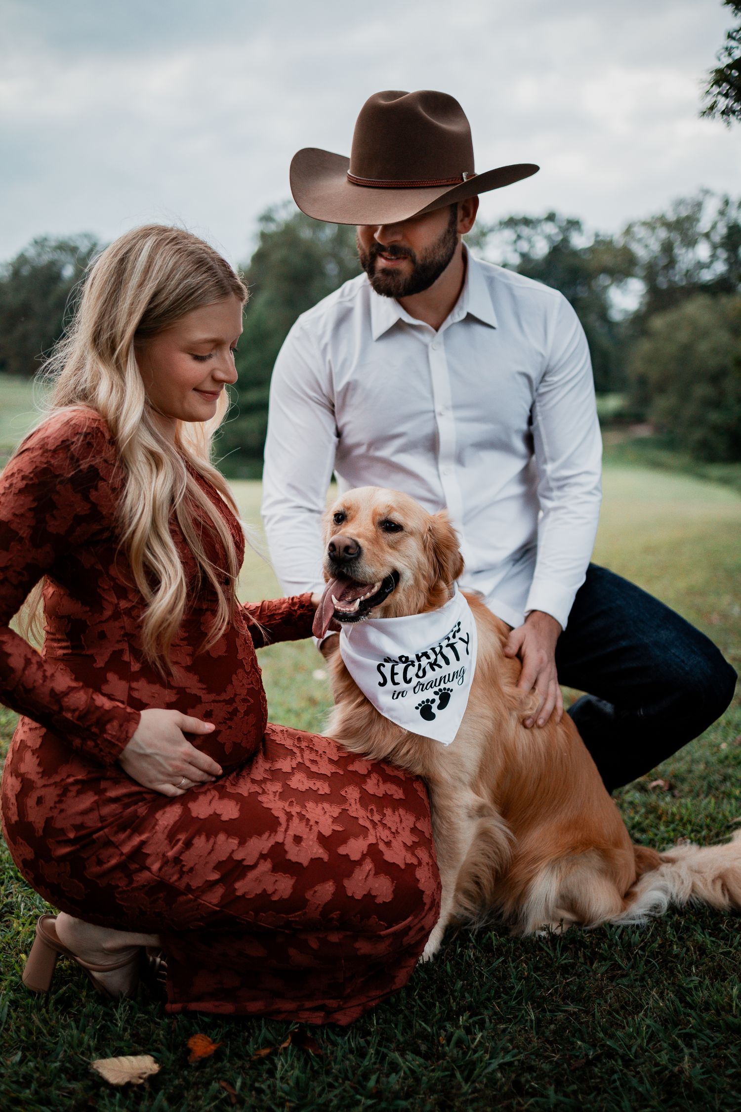 A golden retriever sits with two people on grass wearing a bandana.