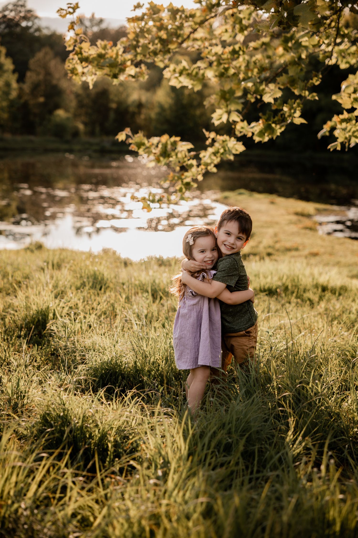 Two young children hugging in a sunlit meadow near a pond, surrounded by lush green grass and golden autumn trees.